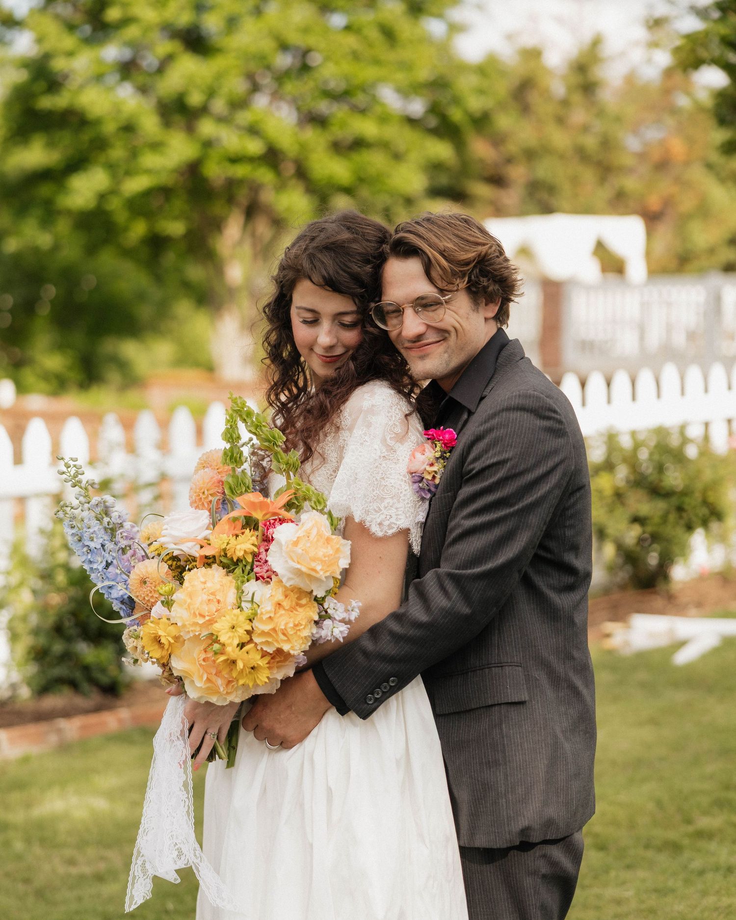 Bride and groom embrace outdoors, she holds colorful bouquet in white lace dress, he wears gray suit by white picket fence.