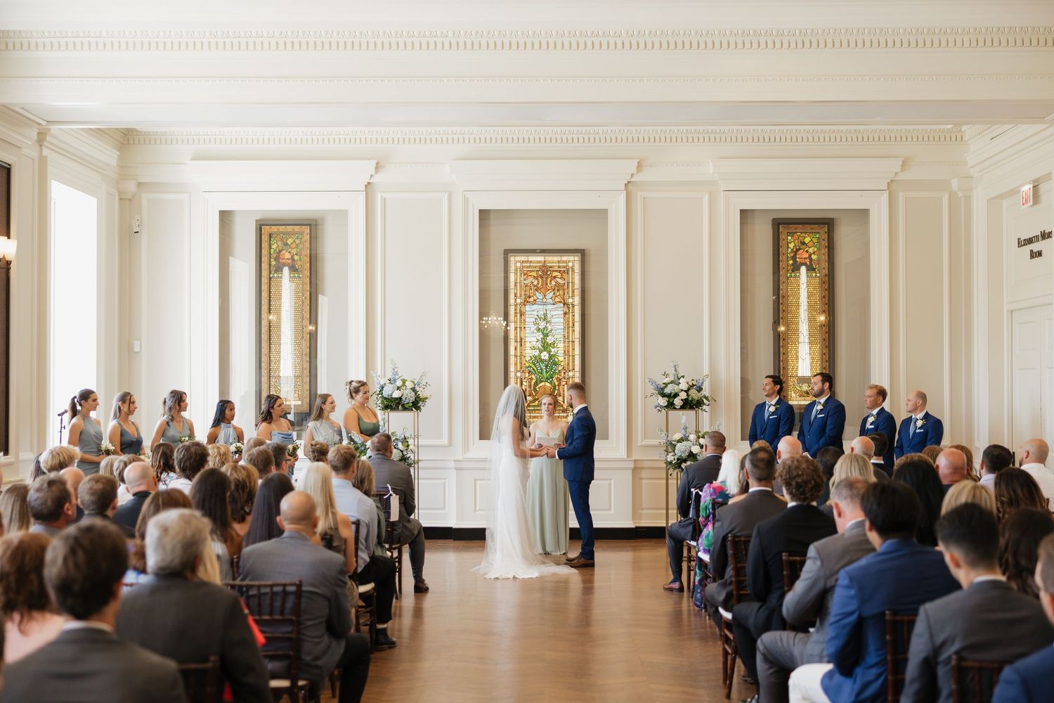 Wedding ceremony taking place in an elegant white hallway with guests seated in rows and decorative artwork on walls.