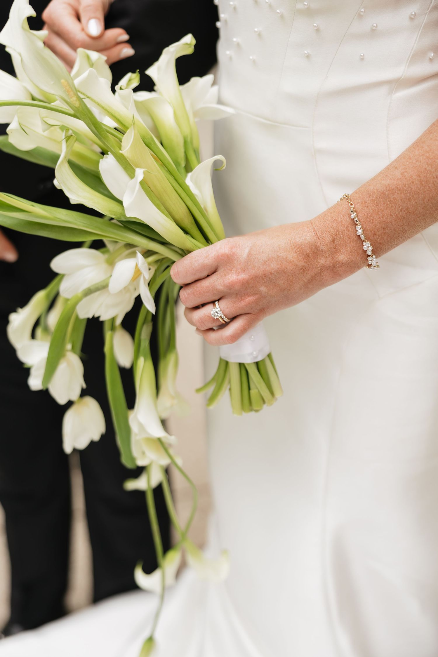Bride holding elegant white calla lily bouquet, wearing diamond ring and bracelet, in white wedding gown.