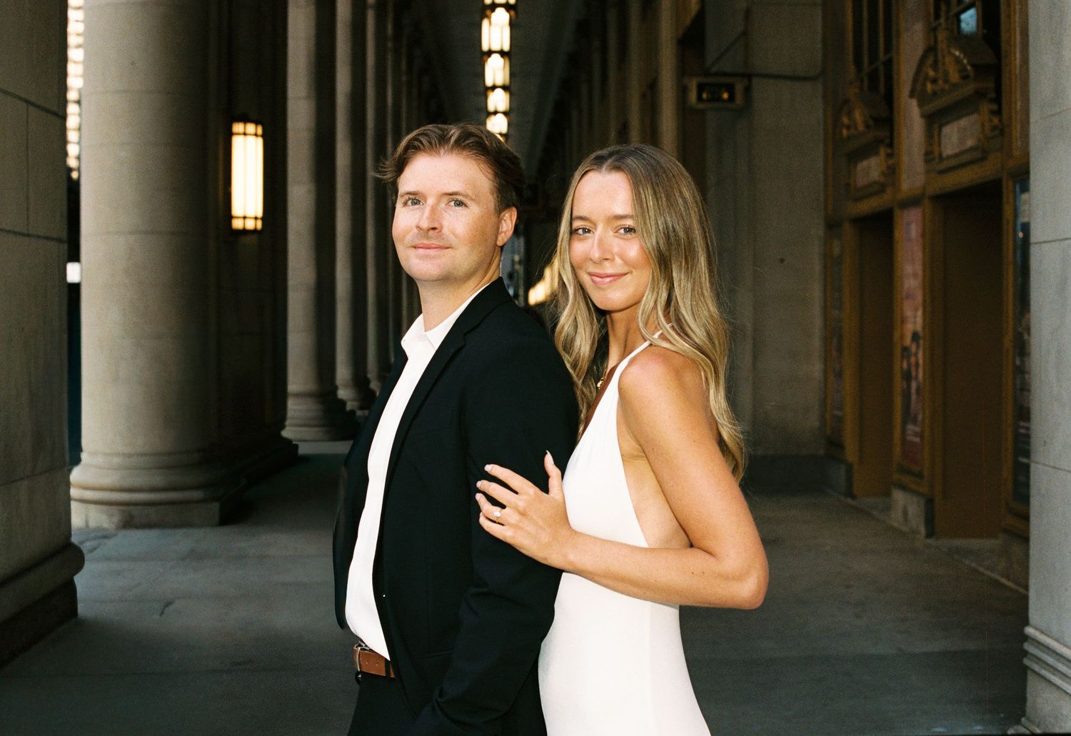 Elegant couple posing in a grand columned hallway, man in black suit and woman in white dress.