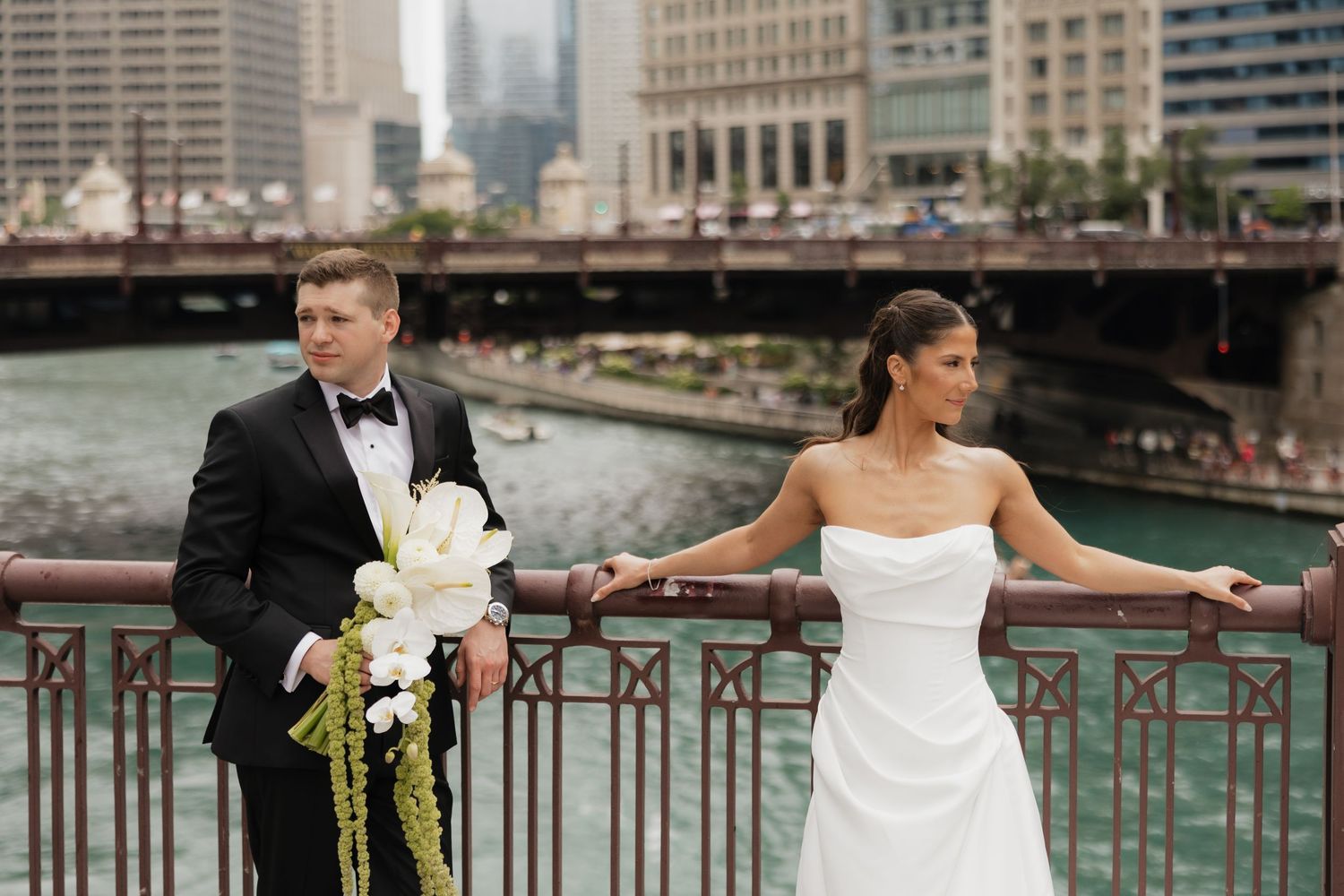 Bride in a strapless white gown and groom in a black tuxedo with a bouquet pose on a bridge railing overlooking a city river.