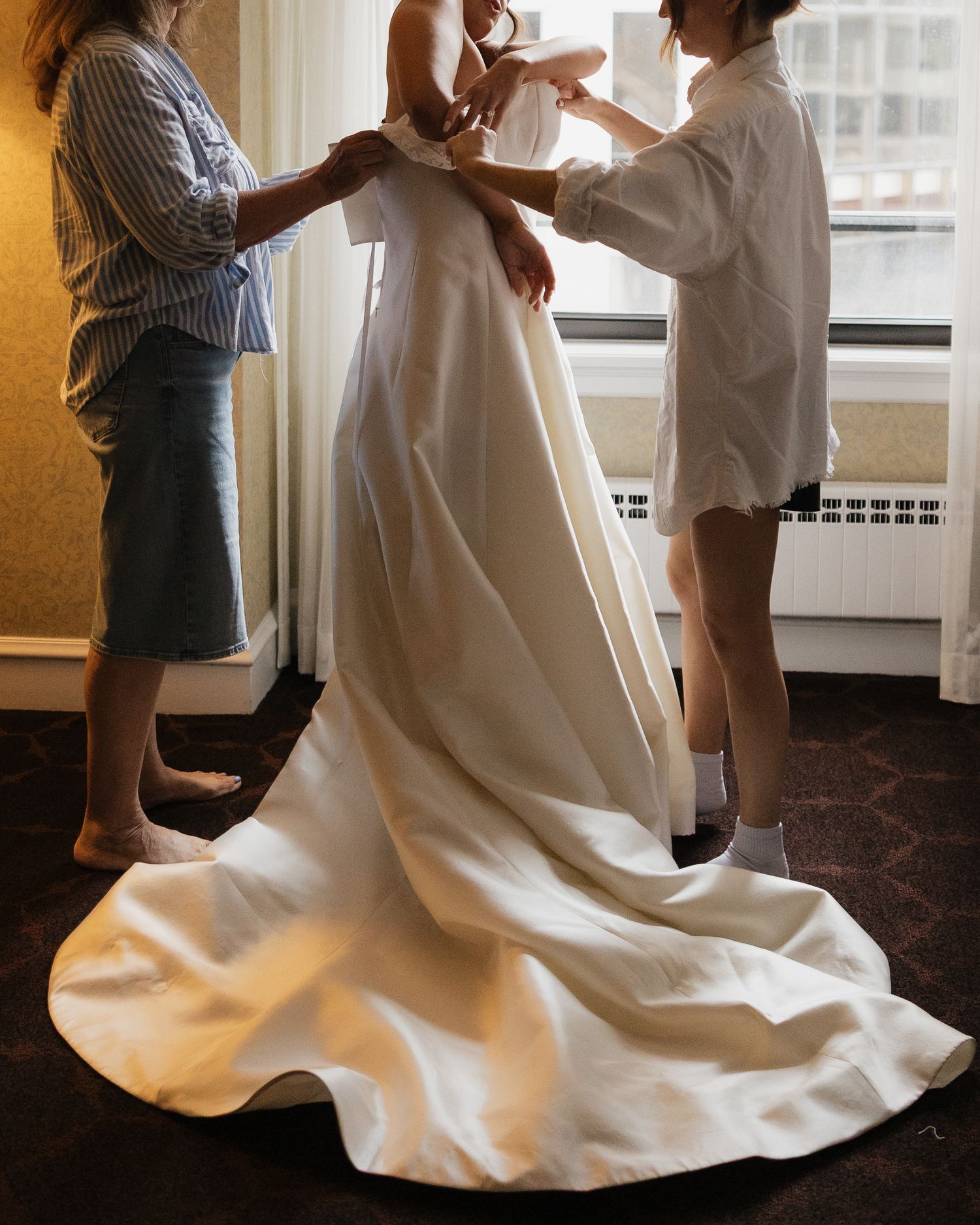 Two women help a bride into her strapless ivory satin wedding gown with a long train in a hotel room.