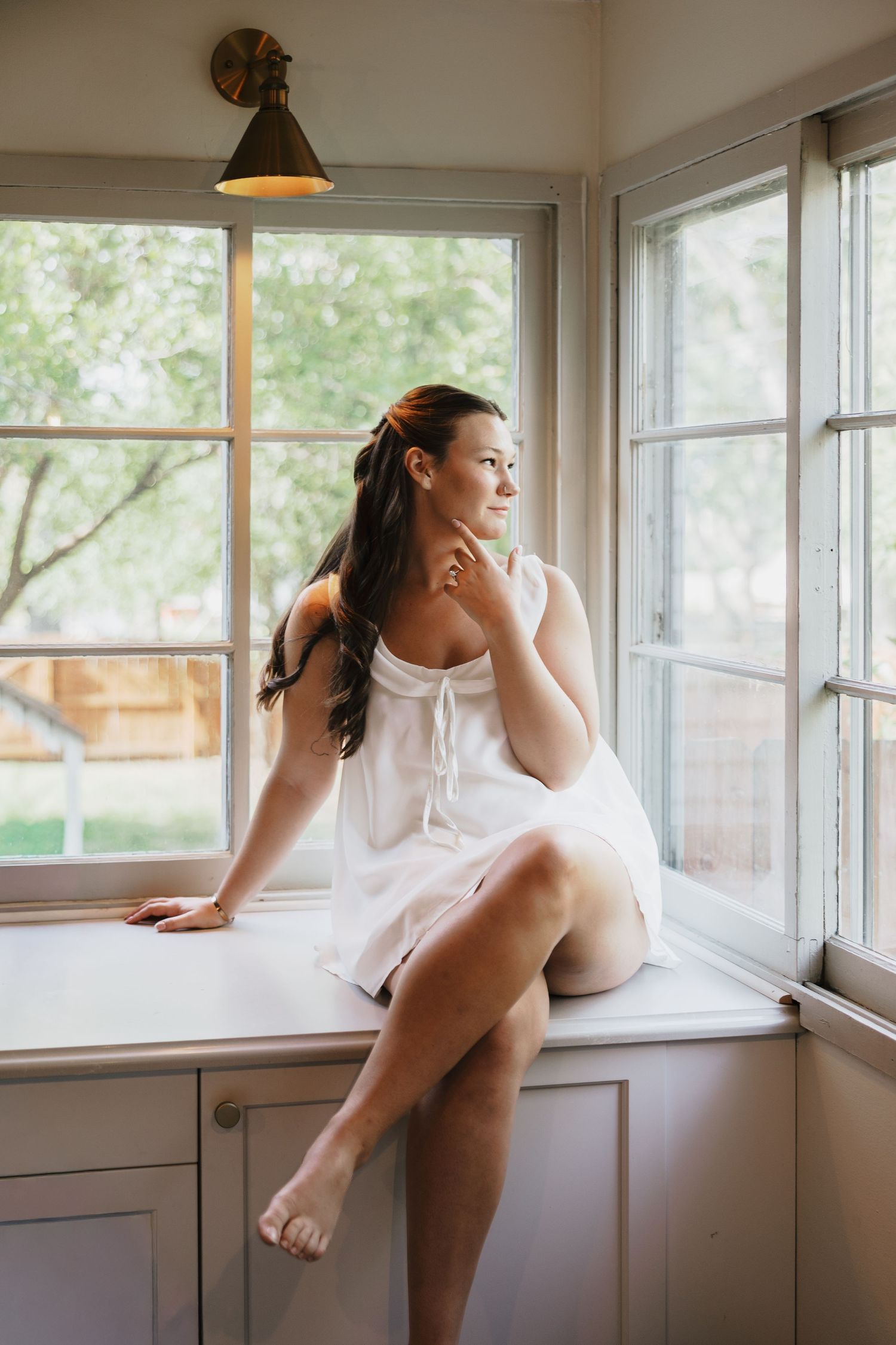 Woman in a white sundress sits on a sunlit window seat, gazing outside through large windows with green trees beyond.