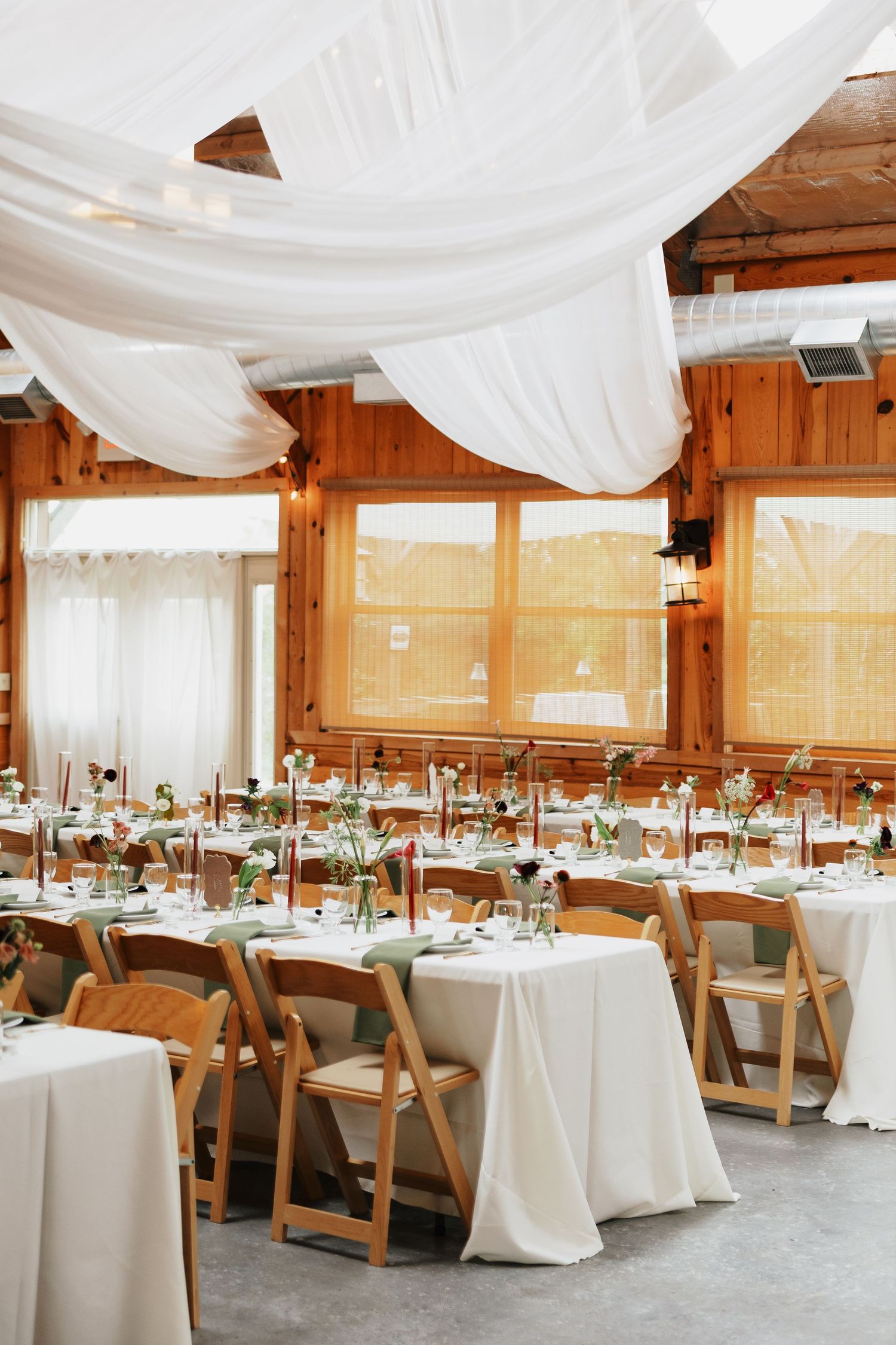 Rustic wooden venue set for a wedding reception with white draped fabric, long tables, linens, greenery centerpieces and folding chairs.