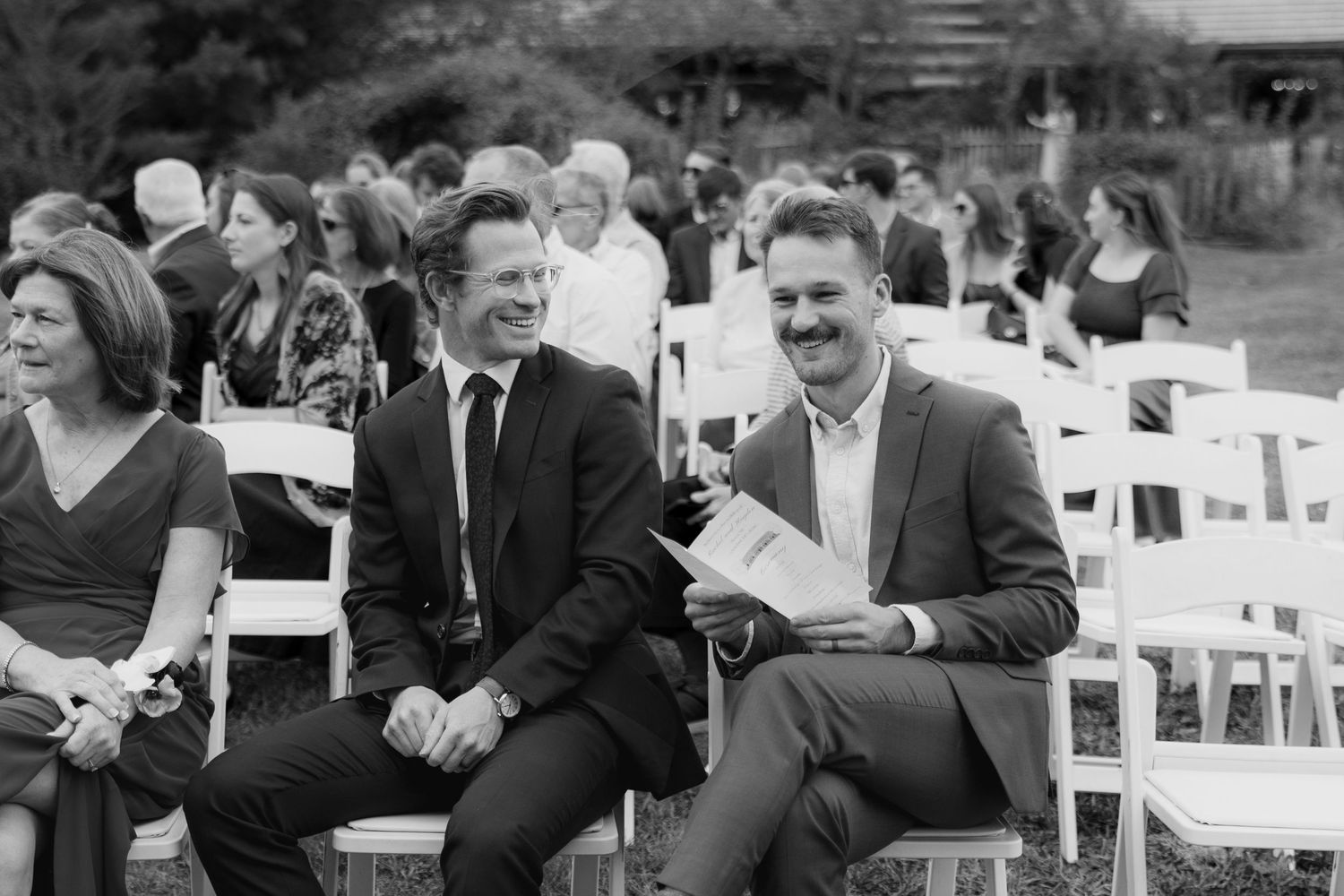 Two smiling men in suits sit on white folding chairs at an outdoor wedding ceremony, with guests seated behind them.