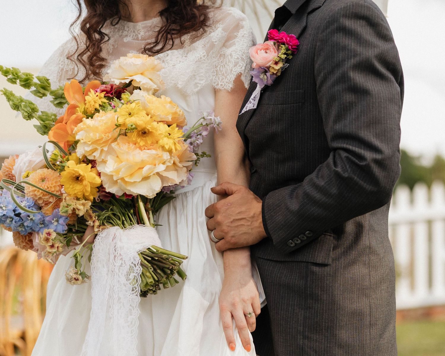 Bride in white lace dress holding colorful bouquet stands beside groom in gray suit at outdoor wedding ceremony.