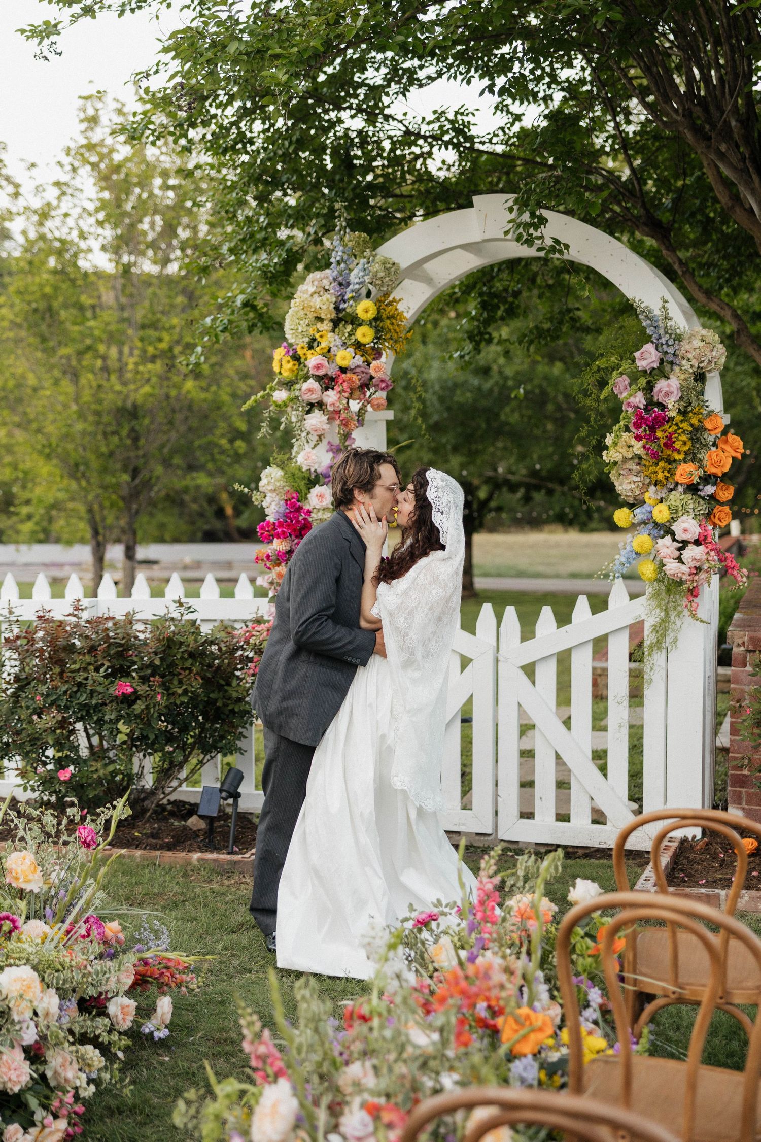 Bride and groom sharing first kiss under colorful floral wedding arch with white picket fence backdrop.