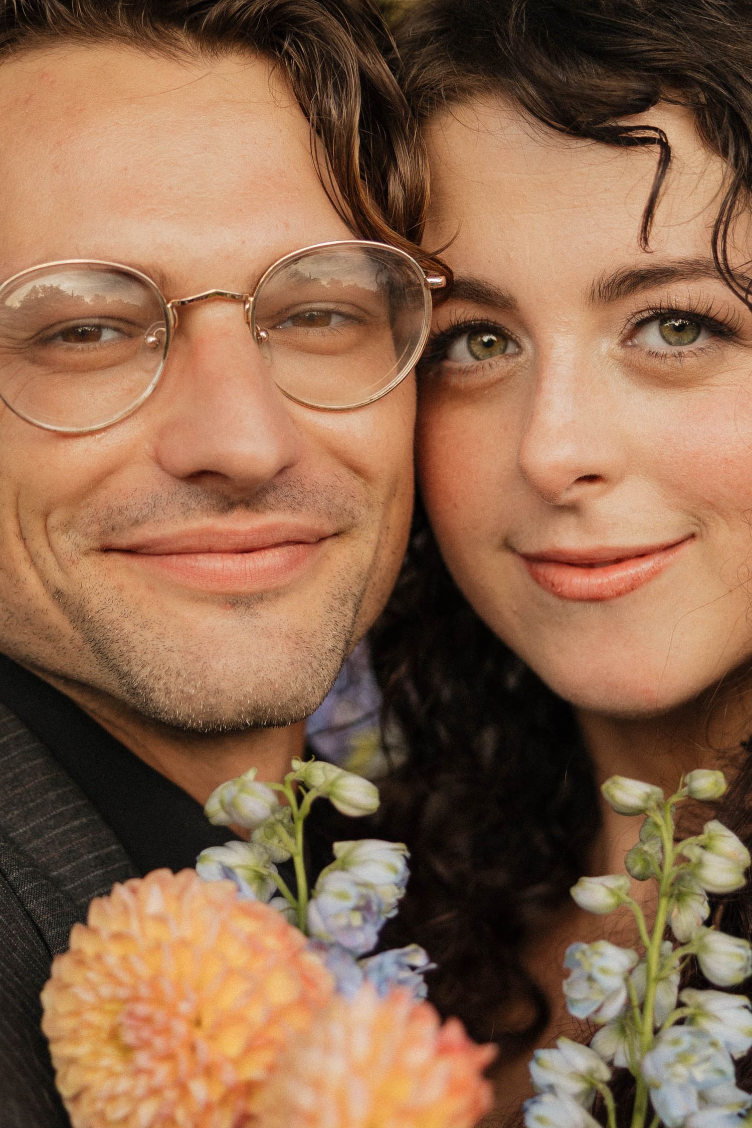 Close-up portrait of a smiling couple with flowers, man wearing round glasses, woman with curly hair.