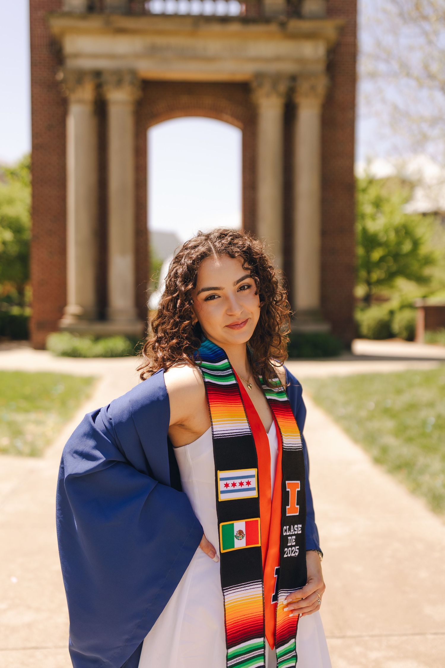 Graduate wearing blue cap and gown with colorful graduation stoles poses for photos at outdoor university colonnade.