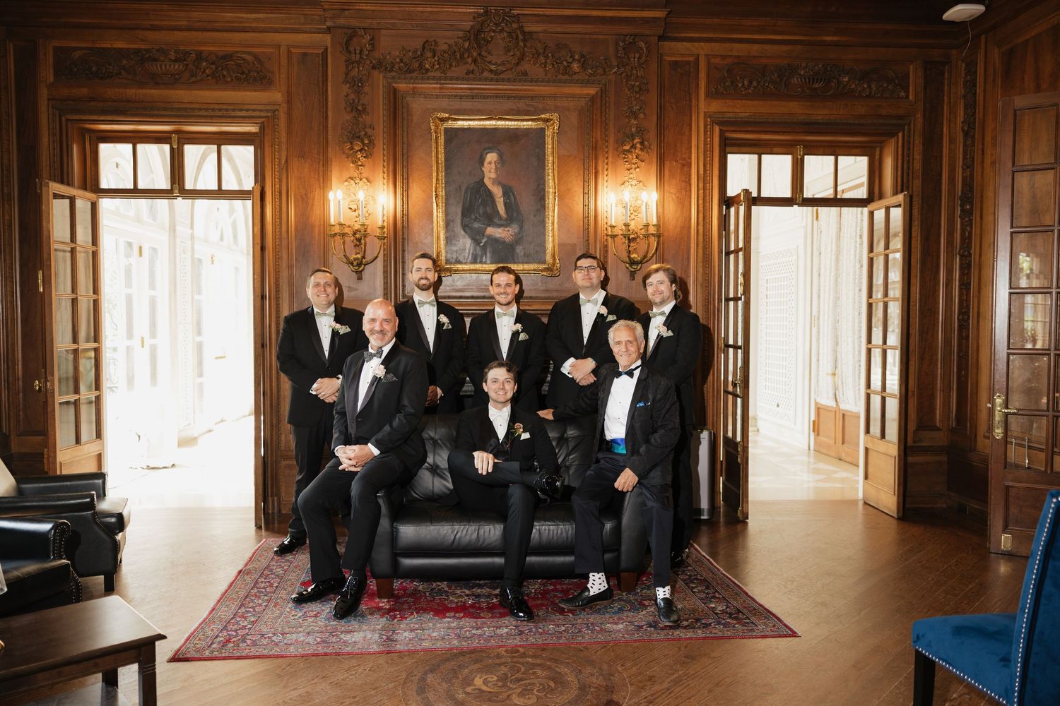 Groomsmen in black tuxedos pose elegantly in a wood-paneled room with classic portrait and ornate decor.