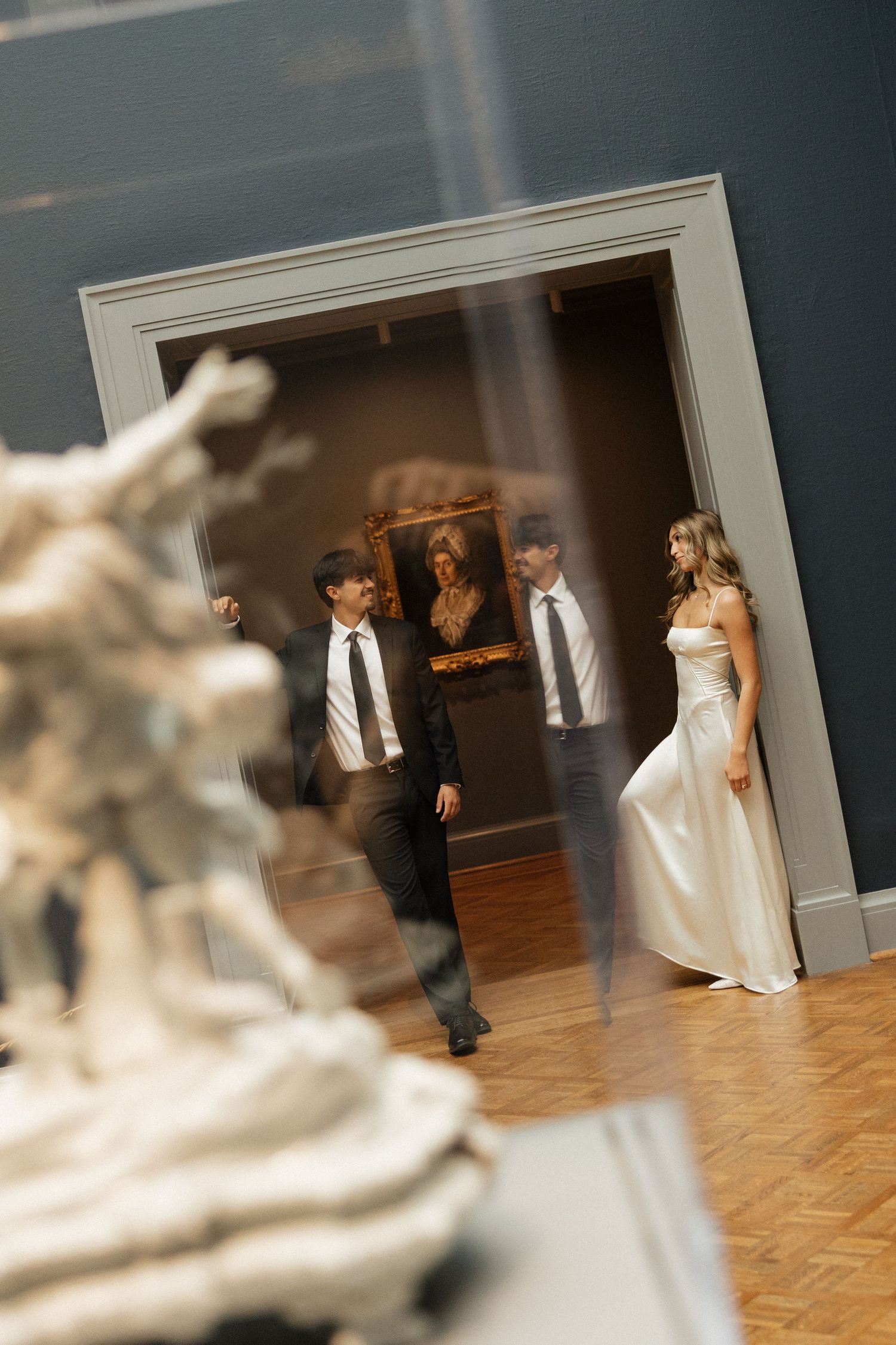 Bride and groom walking through an elegant art museum gallery with sculptures and classic paintings on display.