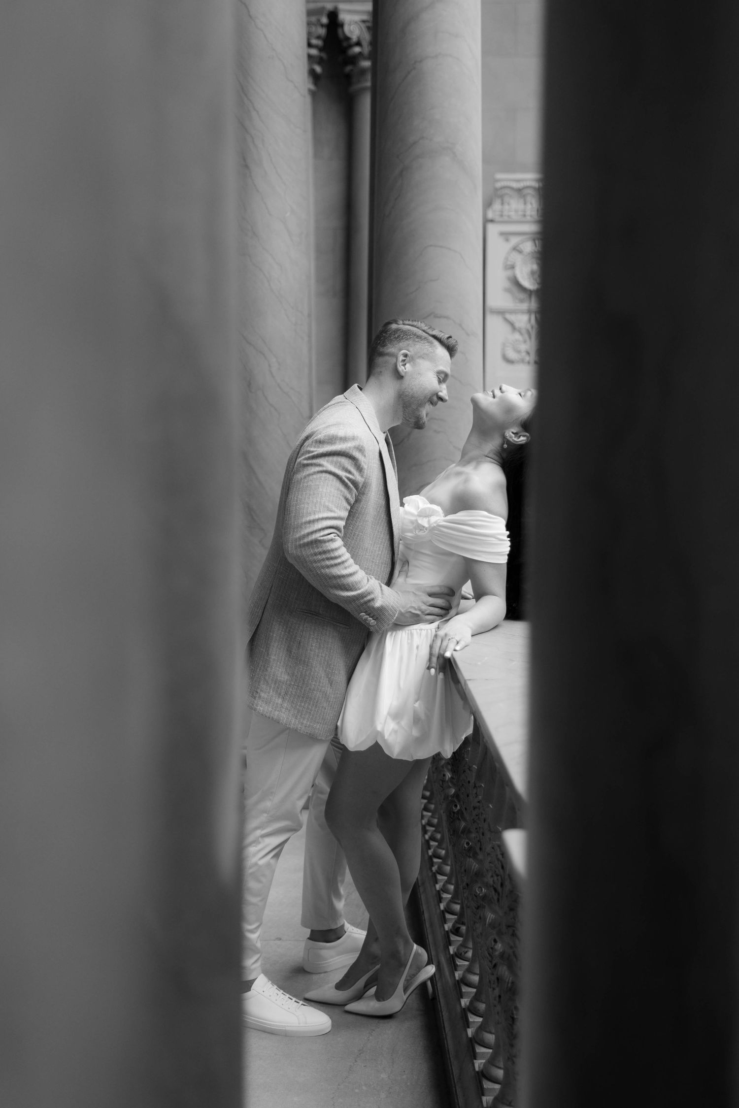 Black and white photo of a laughing couple embracing between ornate stone columns of a classical building.