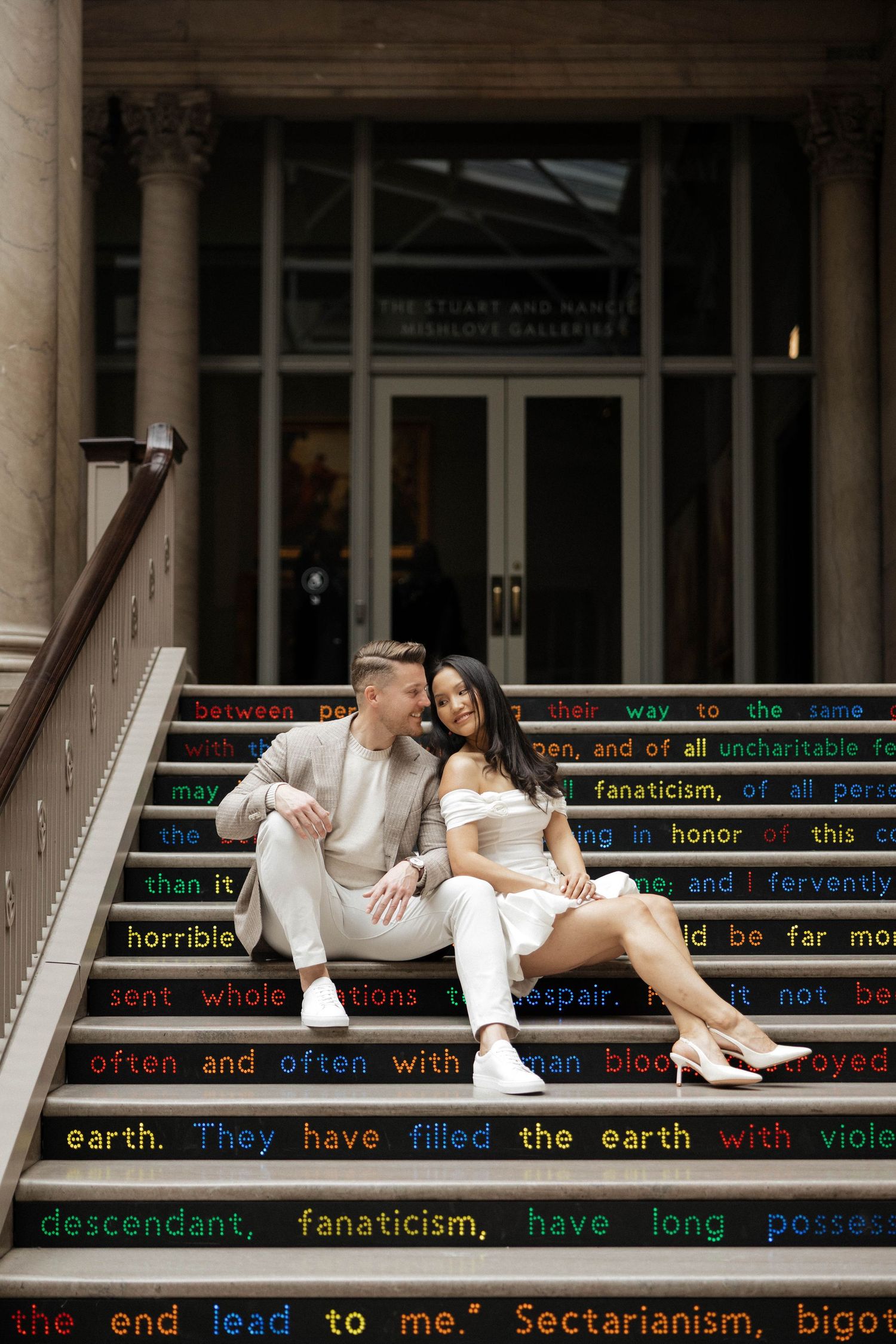 Couple sitting closely on decorated text-inscribed steps outside a gallery building, woman in white dress, man in beige outfit.