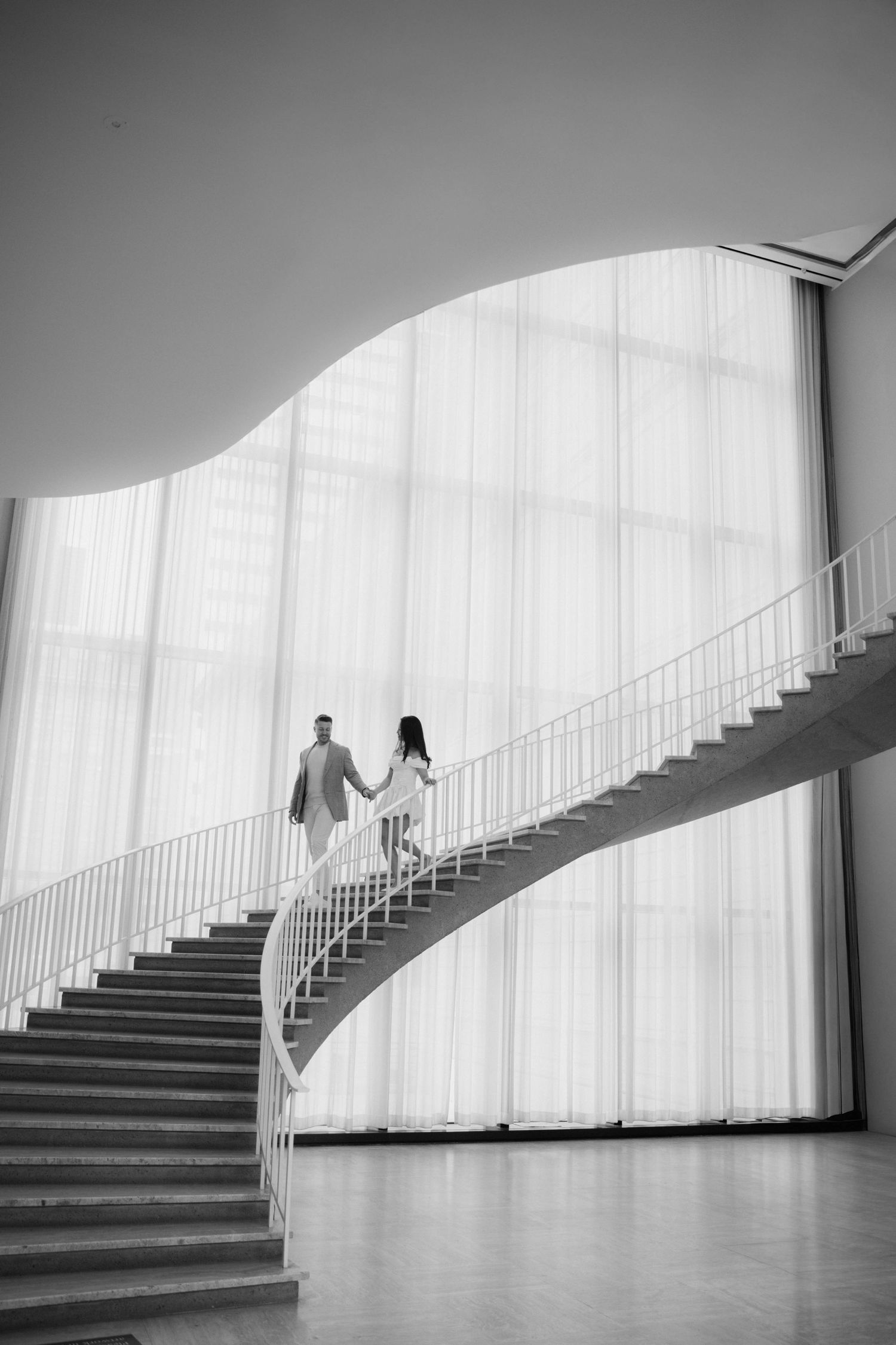 Couple holding hands walking up elegant curved spiral staircase in black and white architectural interior.
