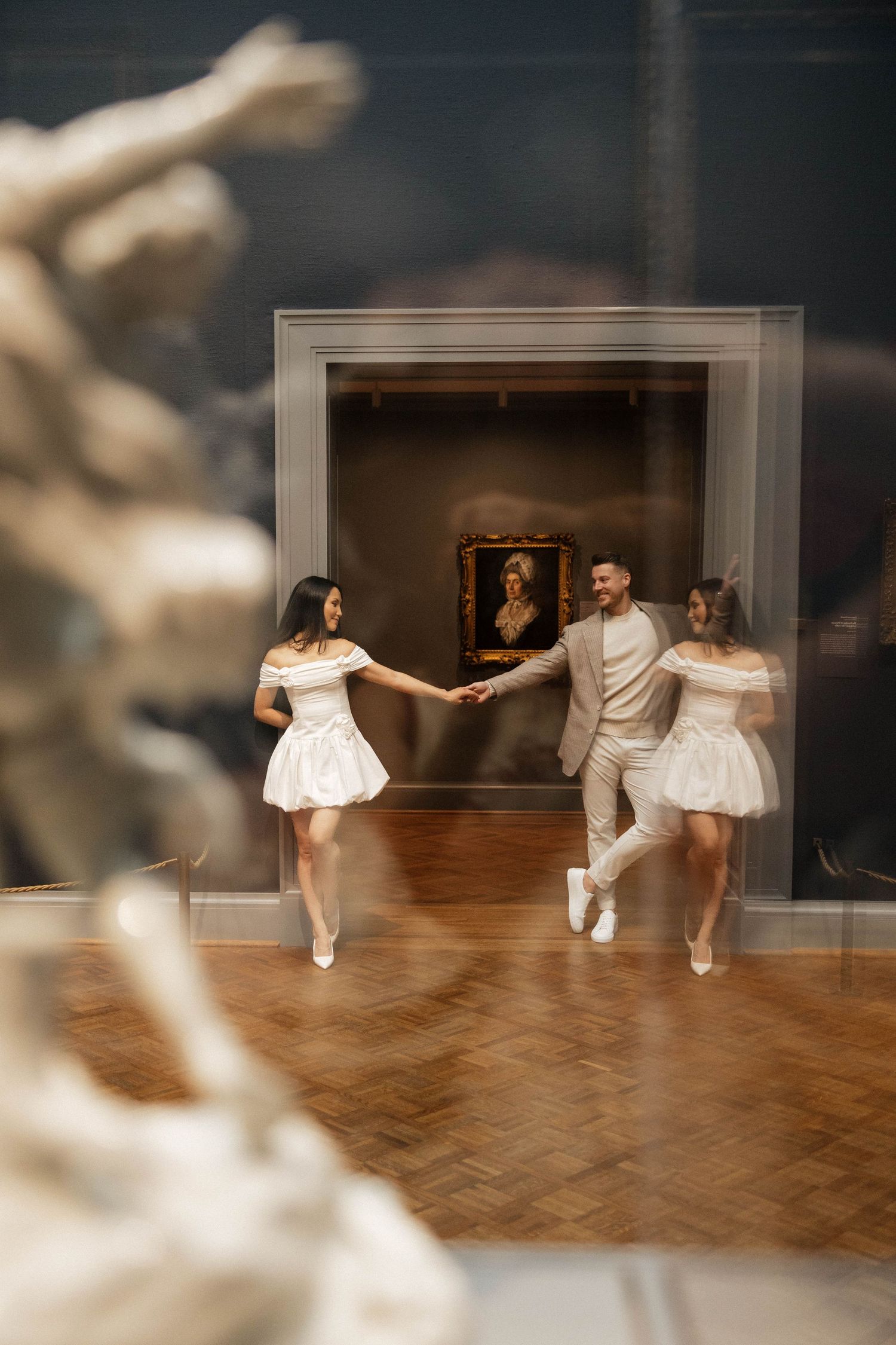 Couple in white outfits dancing joyfully in an art museum, reflected in a mirror beside a classic painting.