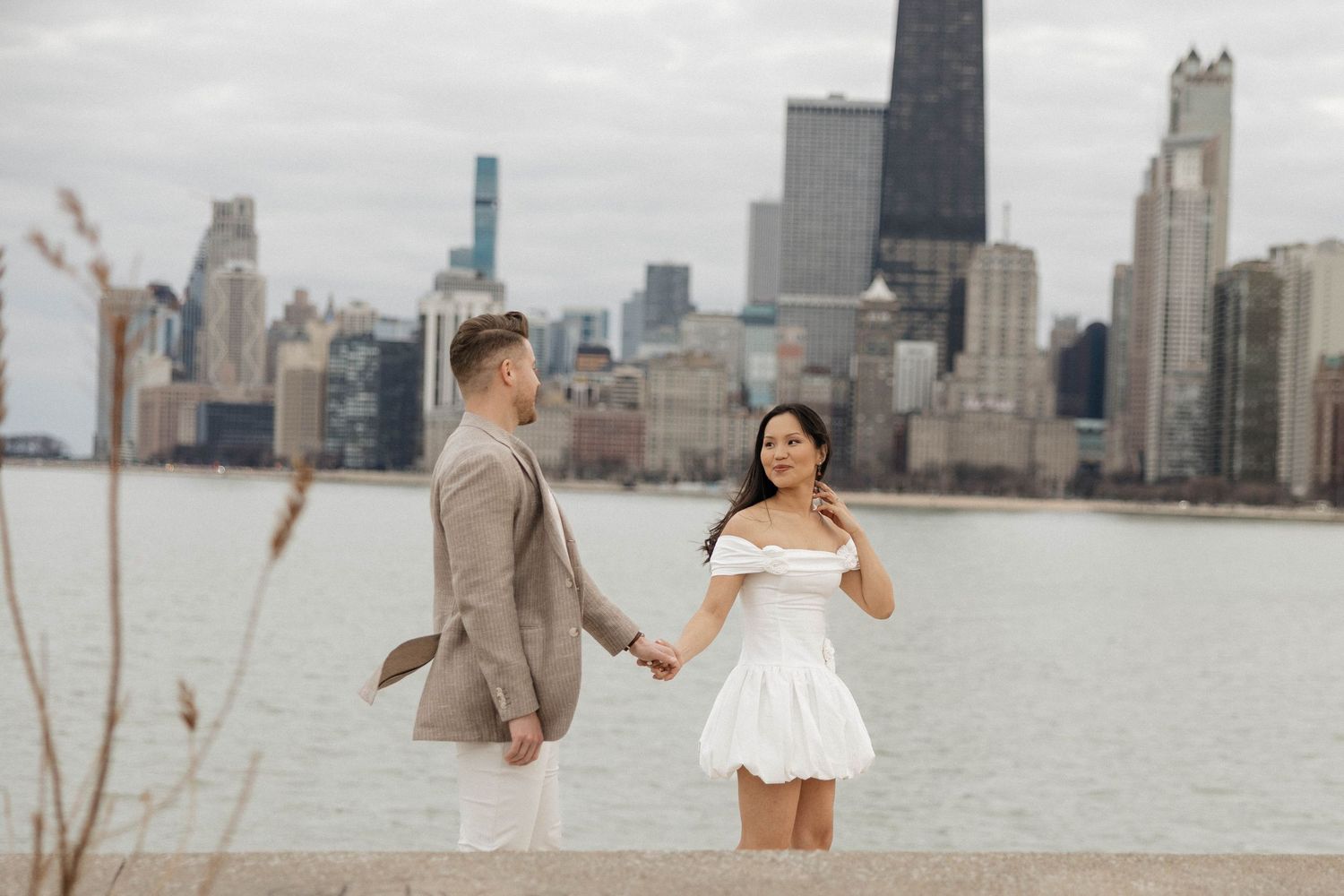 Couple holding hands in front of Chicago skyline, woman in white dress, man in beige suit, near lakefront.