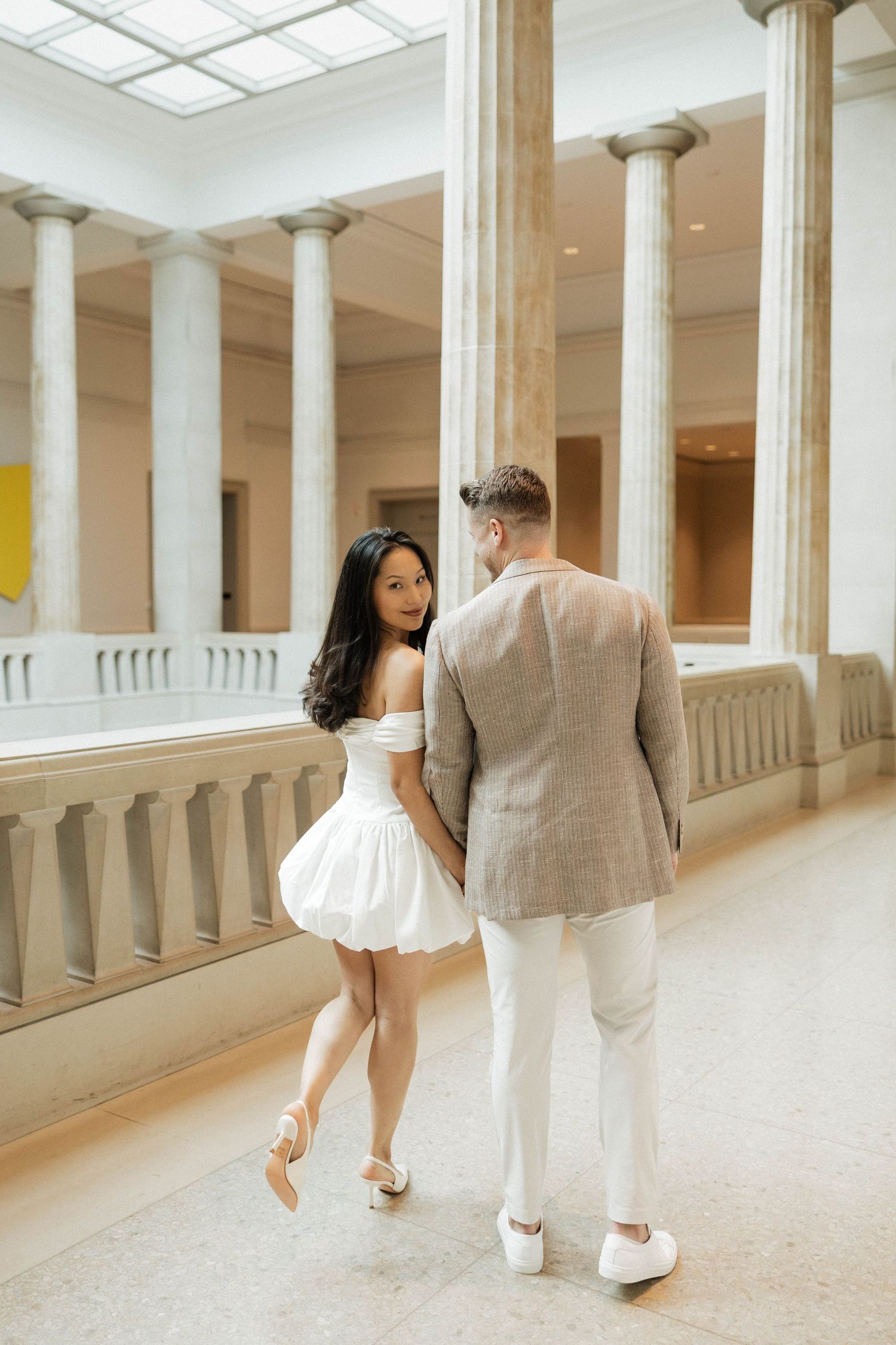 Stylish couple strolling hand in hand in a grand columned gallery, she wears a white off-shoulder dress and heels.