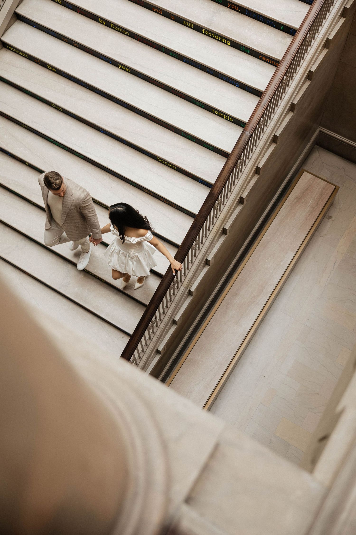 Aerial view of a couple in wedding attire holding hands while walking up grand marble stairs.