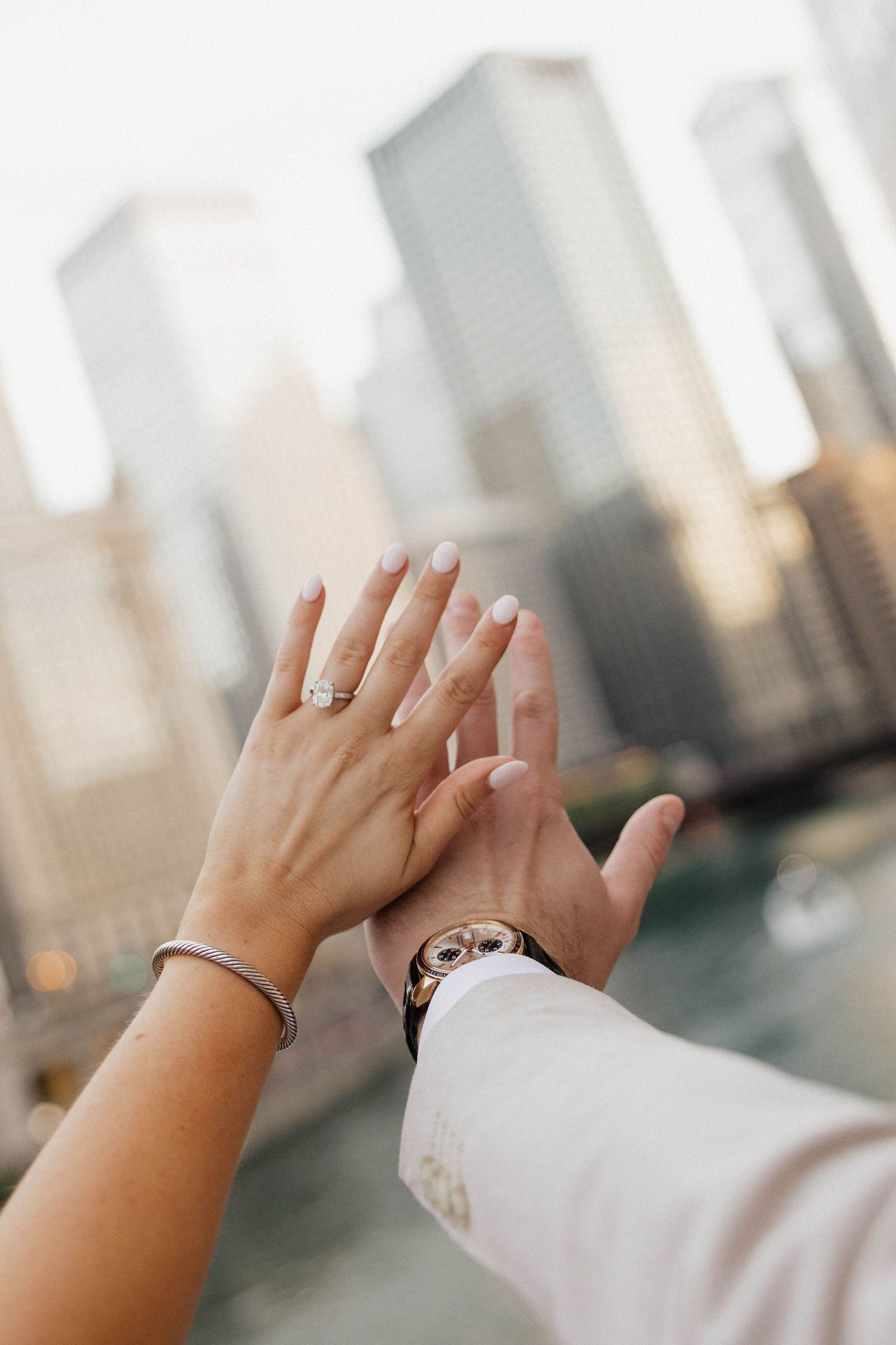 Couple holding hands with engagement ring and watch visible, city skyline with skyscrapers in soft focus background.
