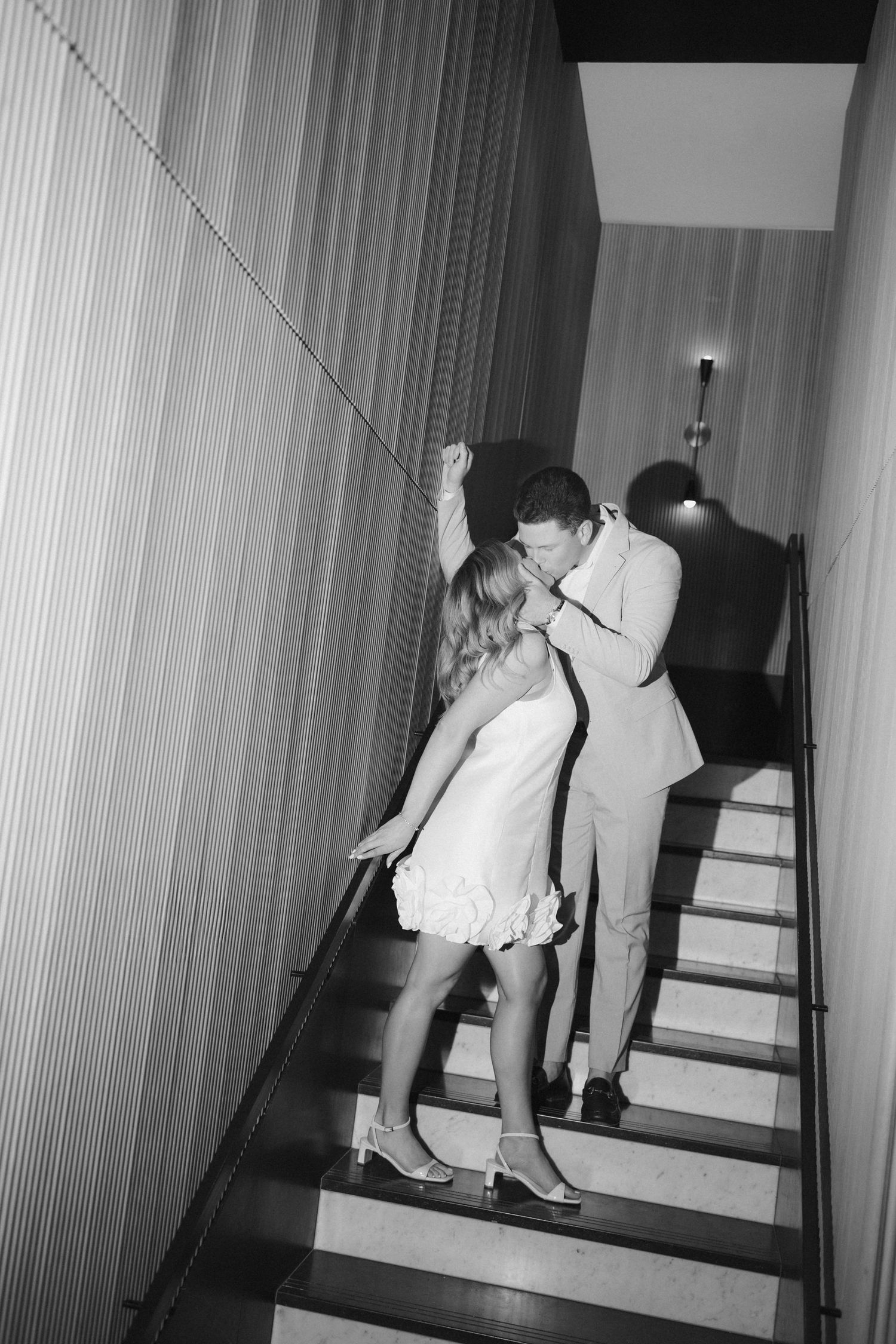 Black and white photo of a couple sharing a kiss on an interior staircase.
