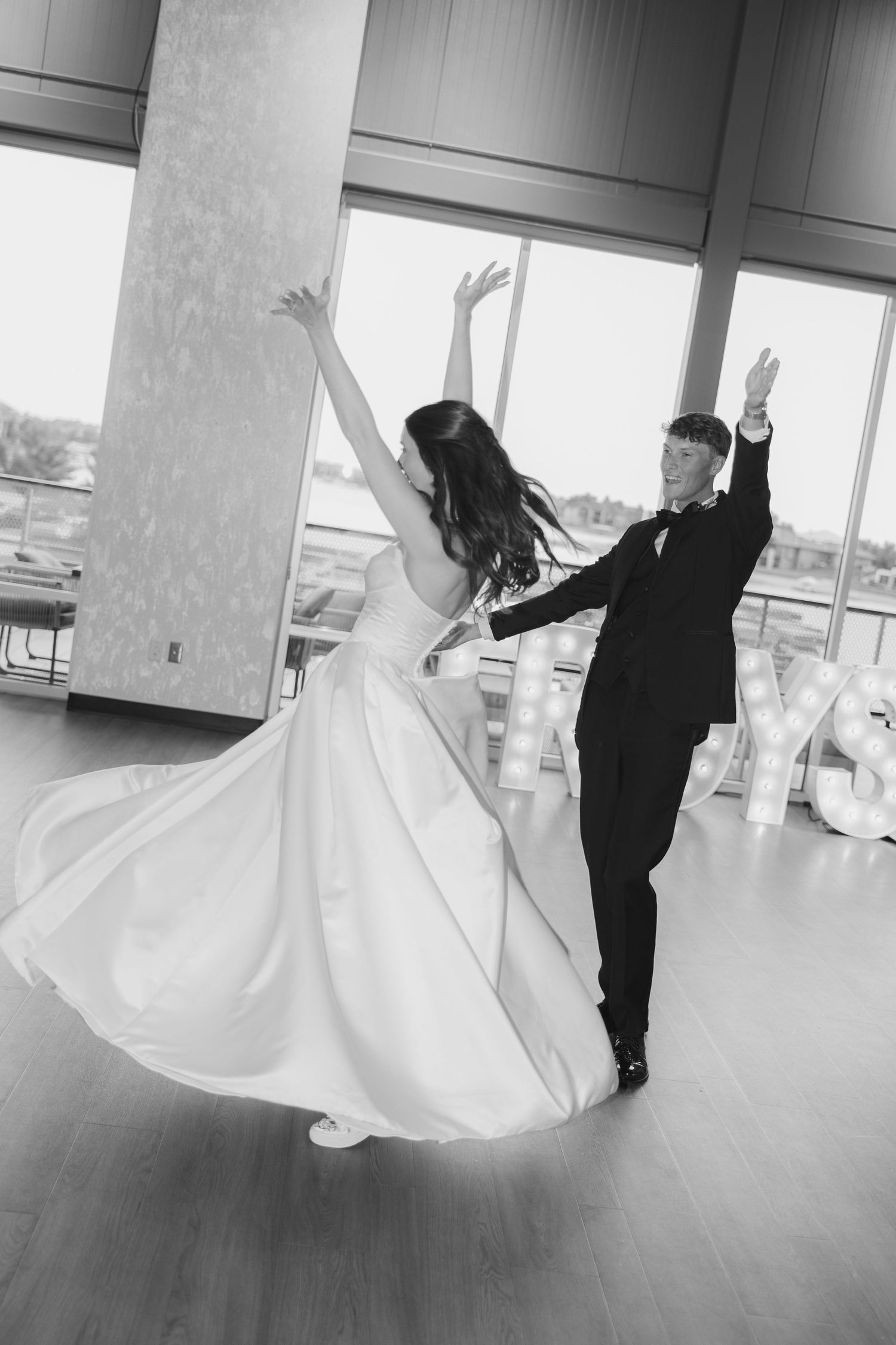 Black and white photo of bride and groom sharing first dance, arms raised gracefully in elegant ballroom.