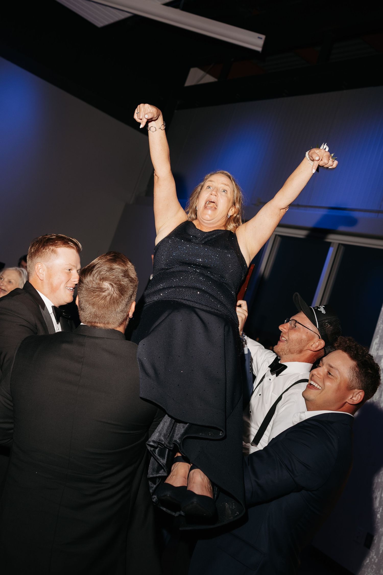 Woman lifted by men in tuxedos celebrating at formal event, arms raised joyfully.