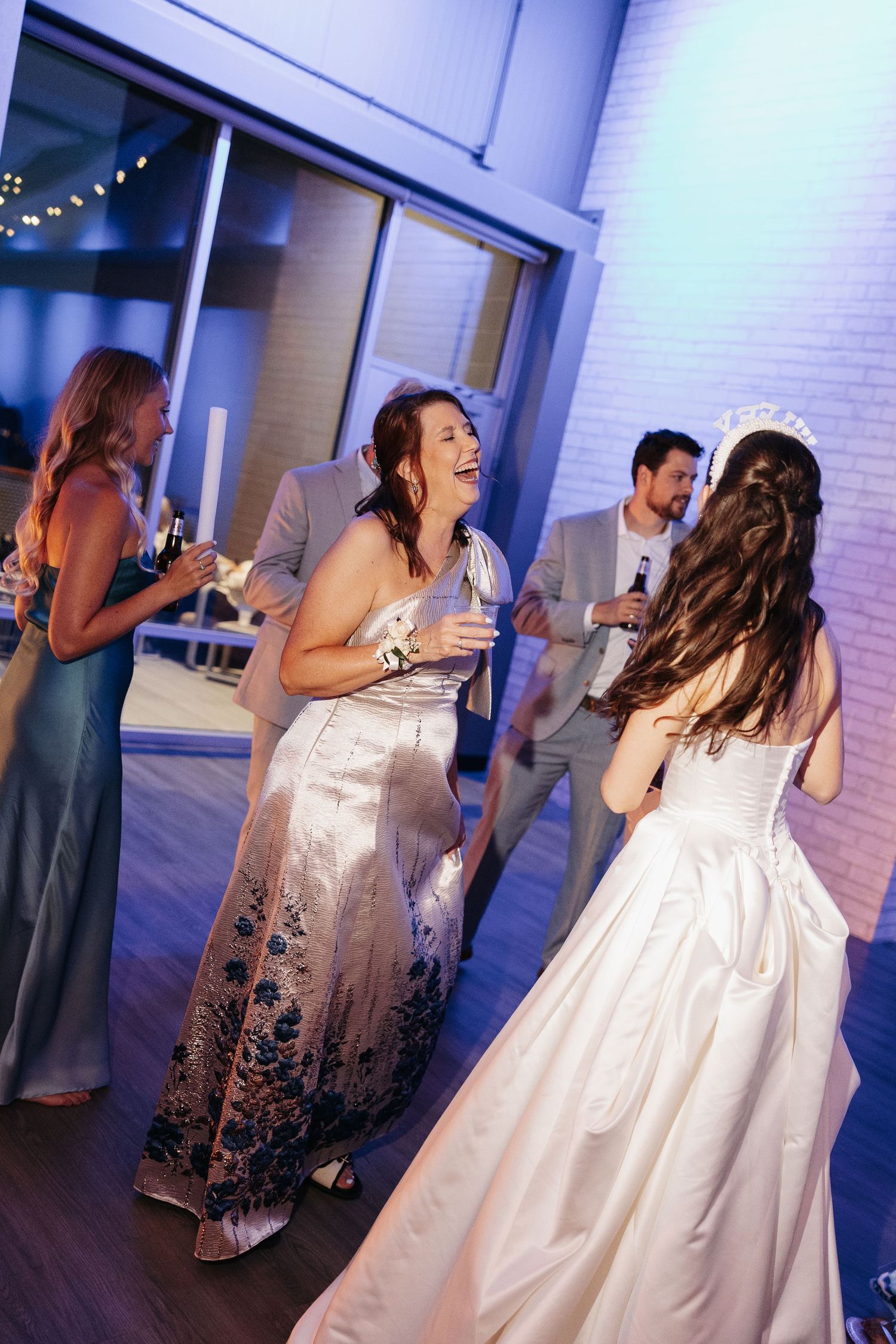 Bride in white gown with tiara laughing with guests in elegant dresses at a wedding reception dance floor.