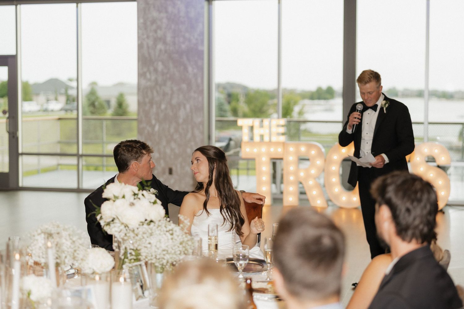 A groom gives a speech at an elegant wedding reception while the bride and groom sit at a decorated head table.