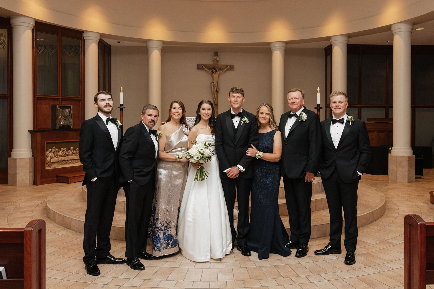 Wedding party poses in church with bride in white gown, groom in tuxedo, and family members in formal attire.