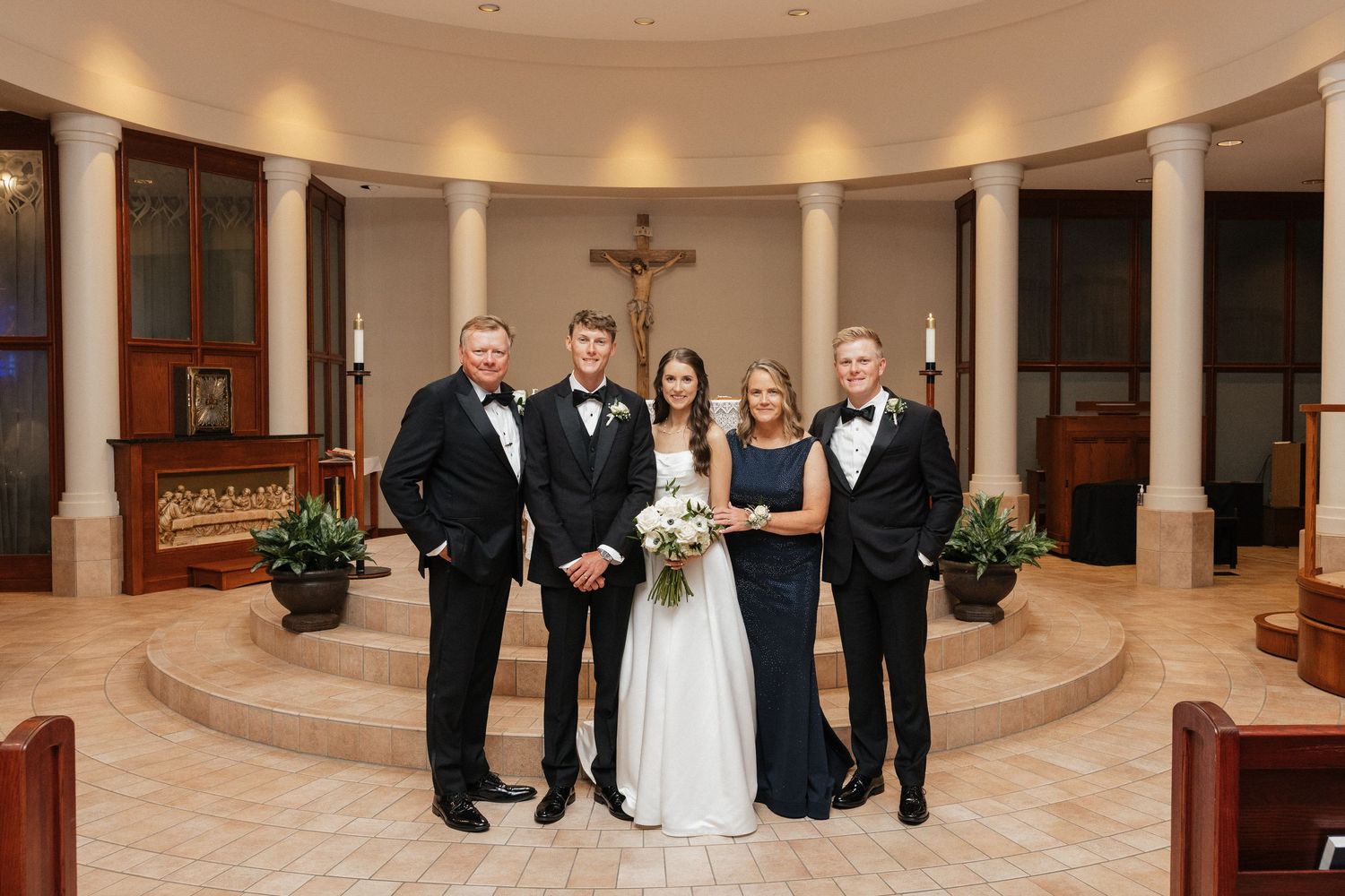Wedding party poses in elegant church with cross backdrop, bride in white gown with bouquet, others in formal black attire.