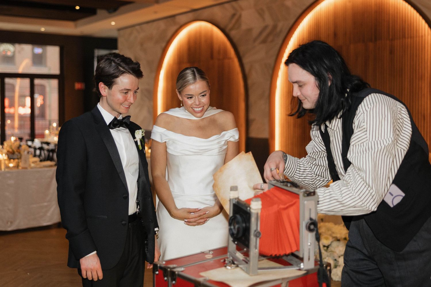 Bride and groom watch a vendor demonstrate a machine at their wedding reception in a warmly lit venue.