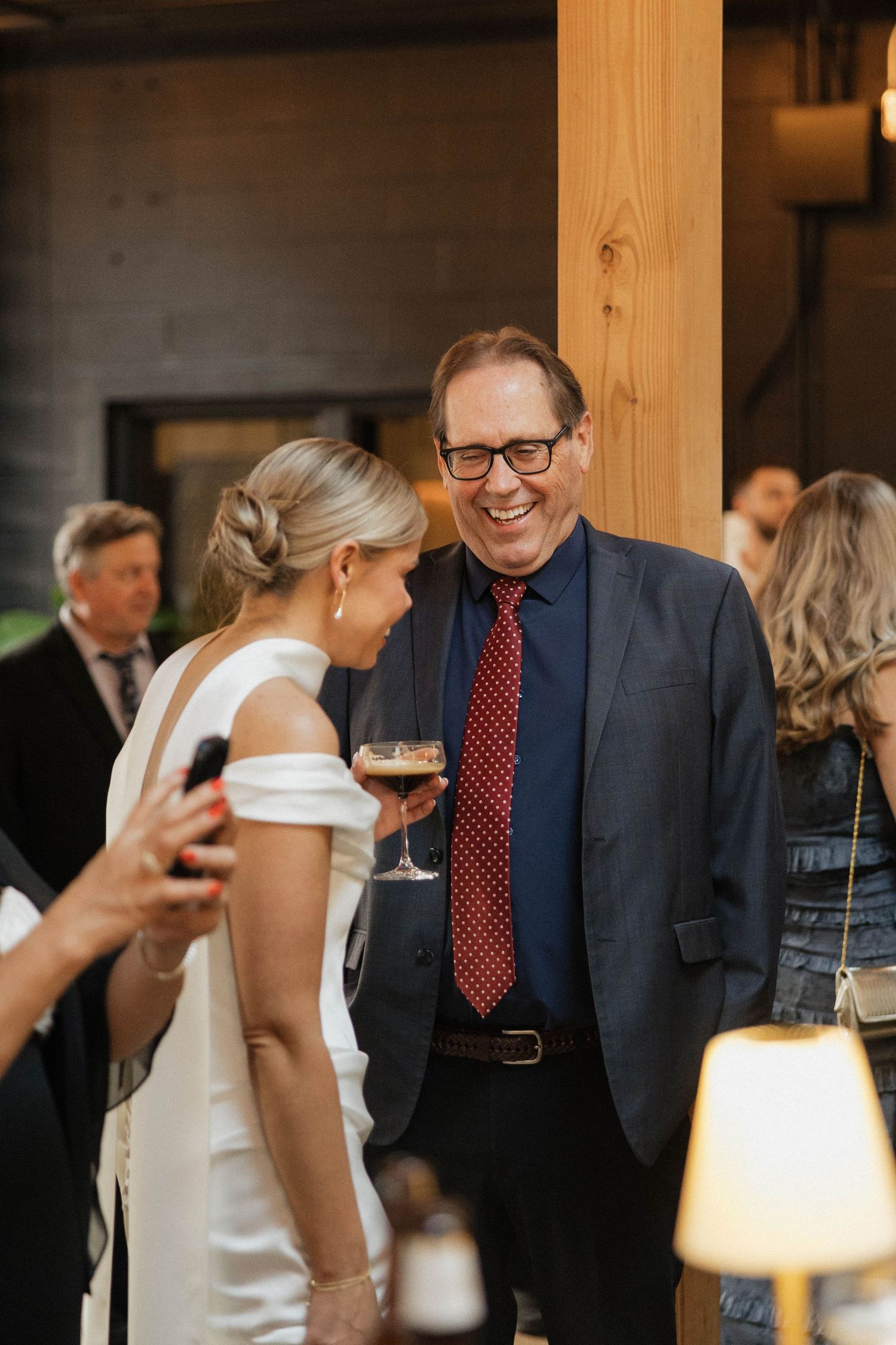 Bride in white gown laughing with older male guest in navy suit and red tie at wedding reception.
