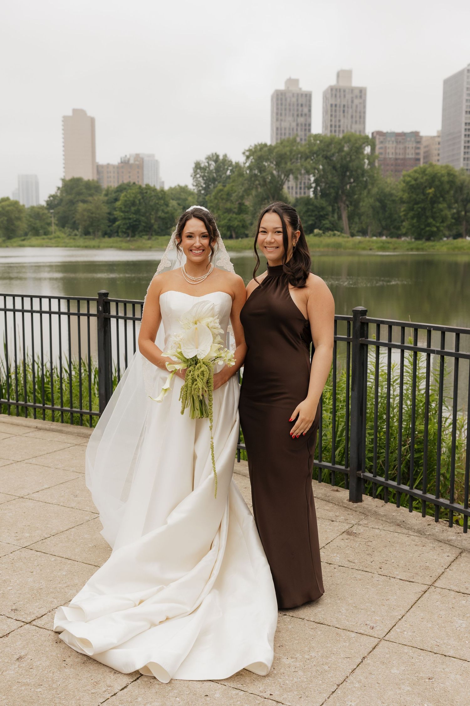 Bride in white gown with veil and bouquet poses with bridesmaid in brown dress by Central Park lake, NYC skyline behind.
