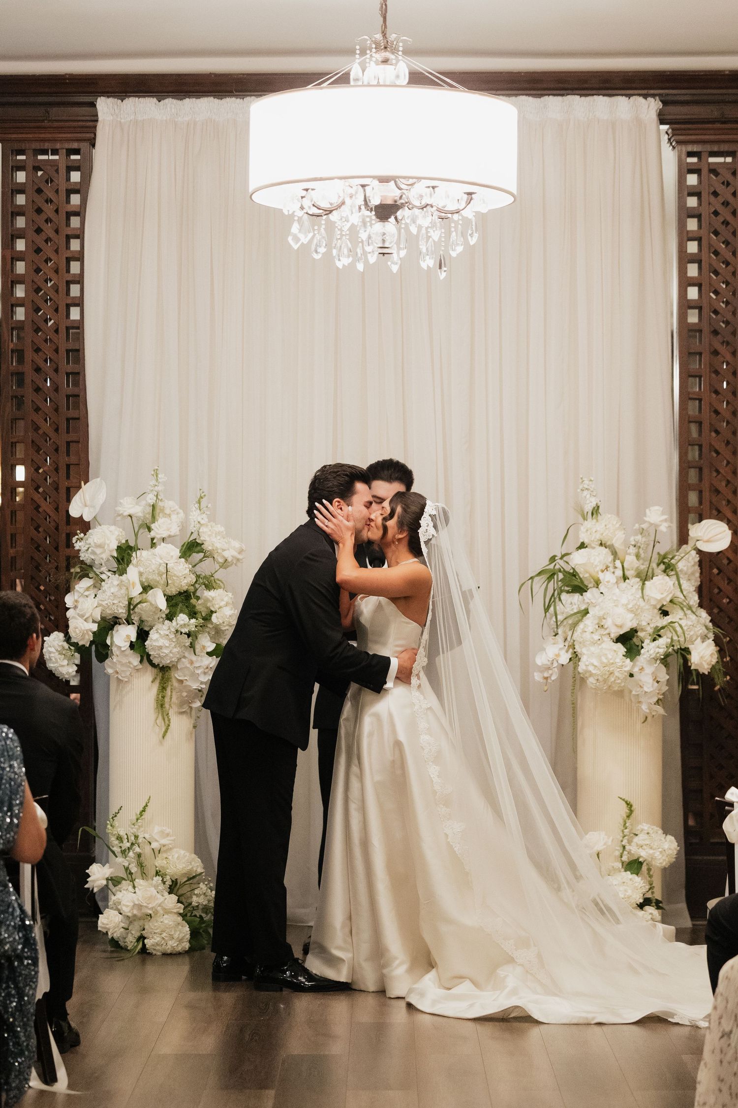 Bride and groom share first kiss at elegant wedding ceremony with white floral arrangements and crystal chandelier backdrop.