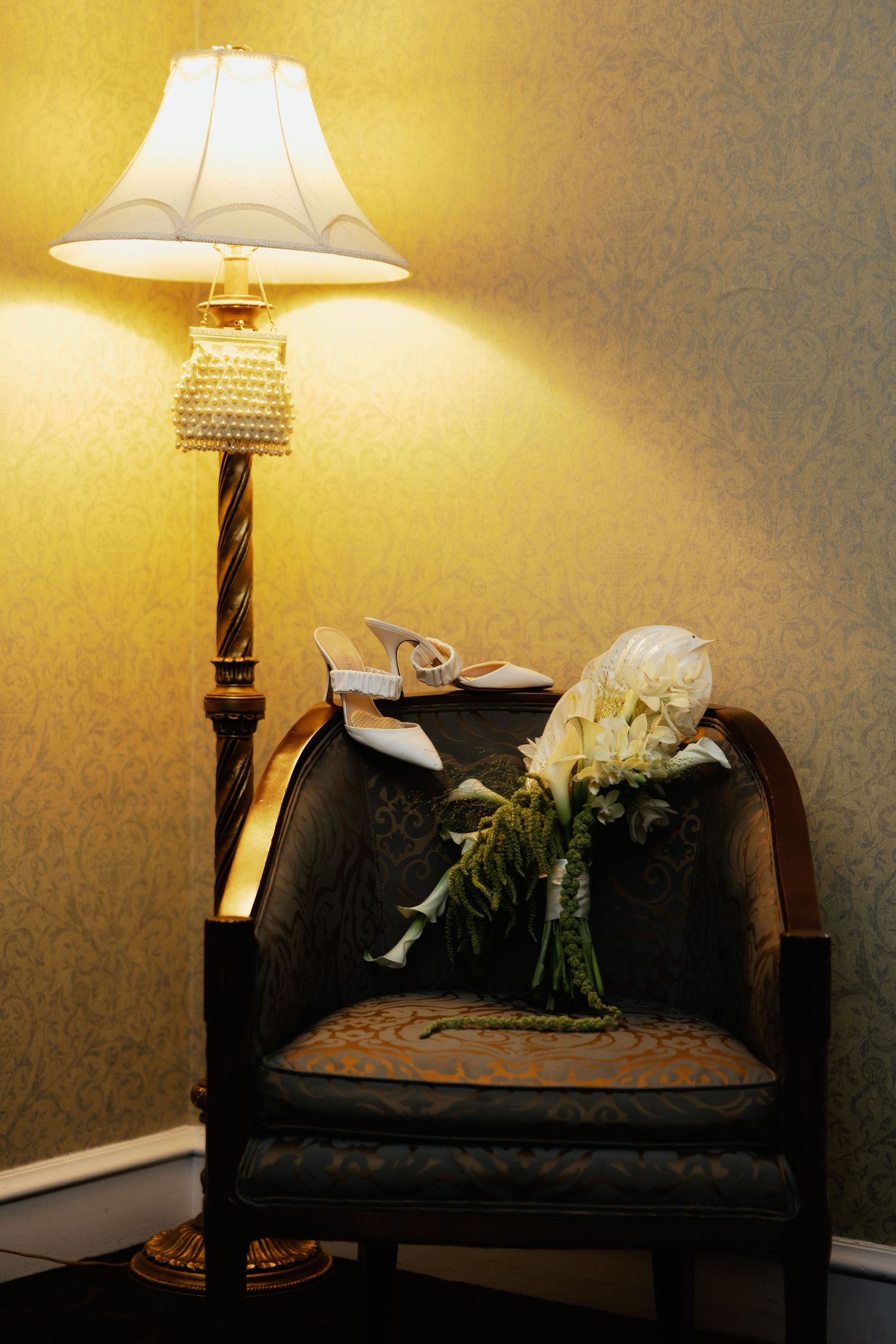 Elegant floor lamp illuminating a vintage chair with bridal bouquet and white heels in a warmly lit corner.