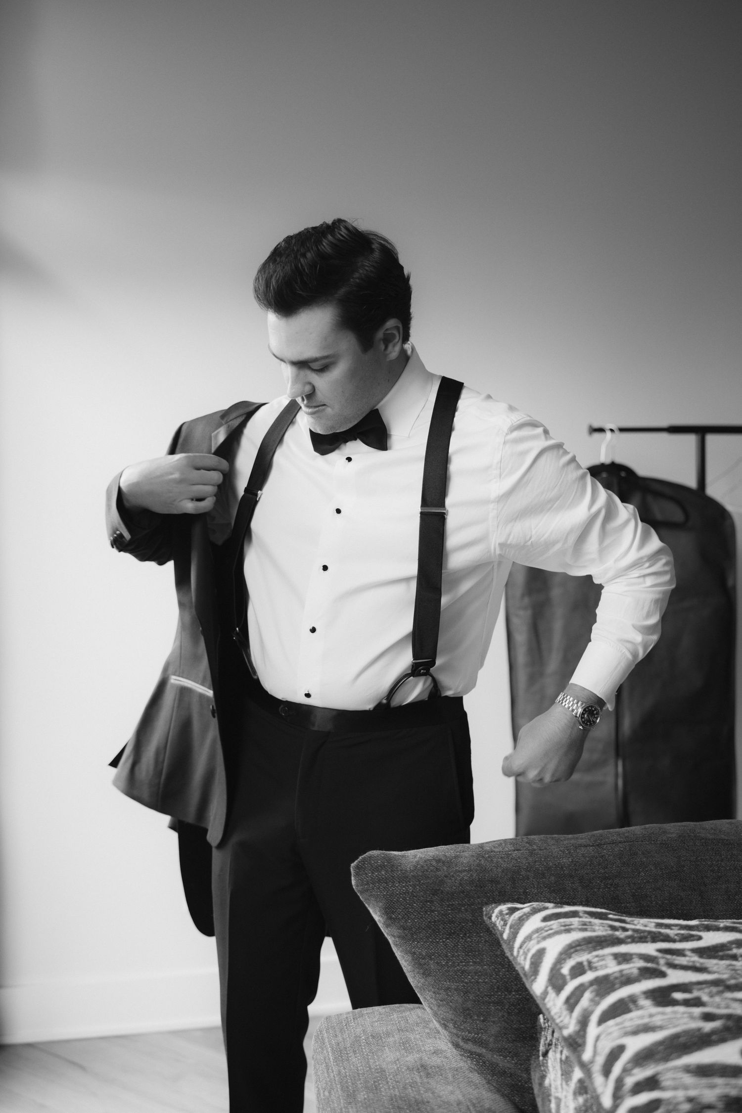 Black and white photo of a groom putting on his tuxedo jacket with suspenders and bow tie before wedding.
