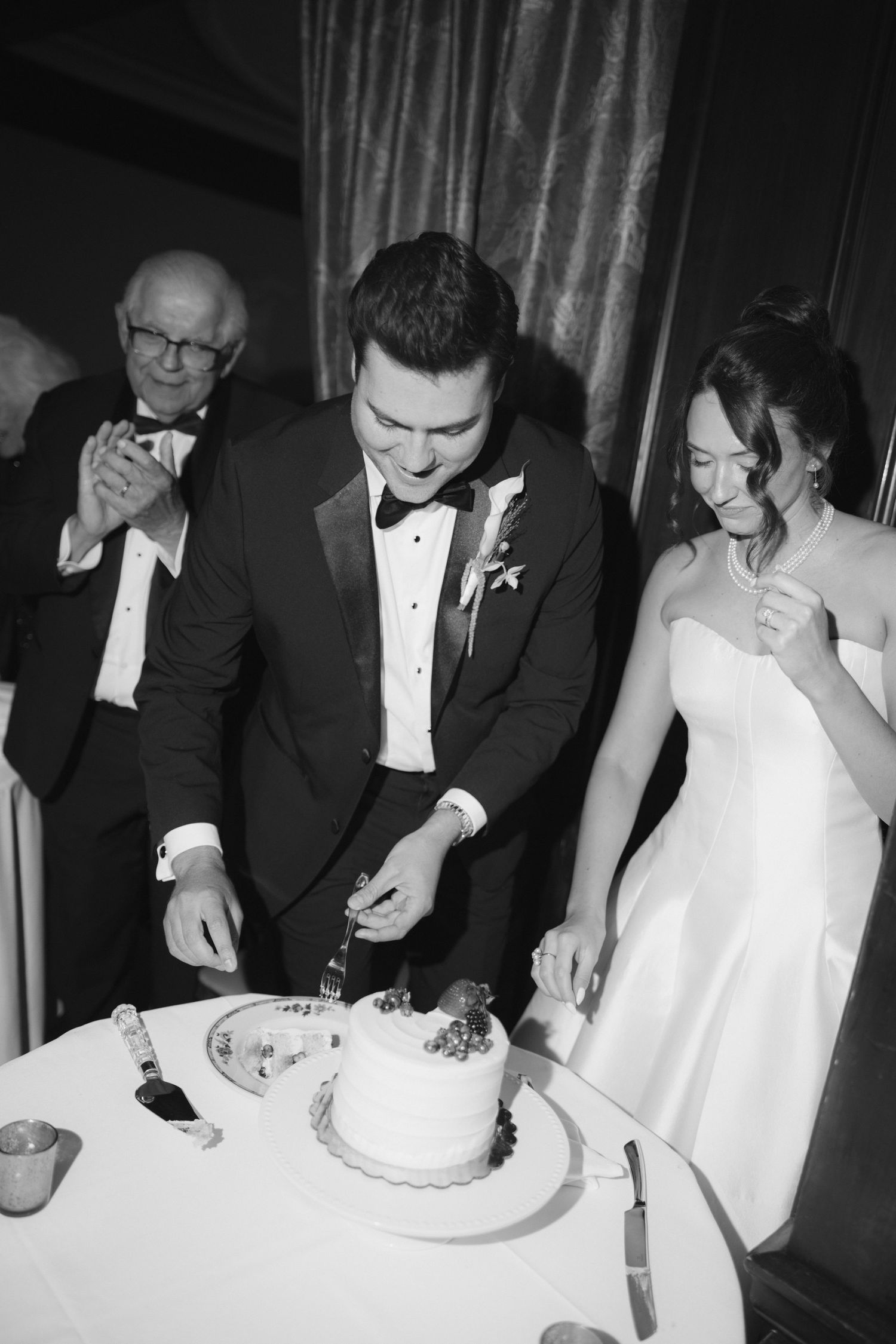 Black and white photo of bride and groom cutting wedding cake together while older guest claps in background.