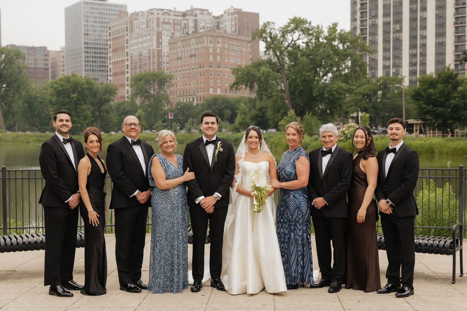 Wedding party poses outdoors near a city park, bride in white gown flanked by family in formal attire.
