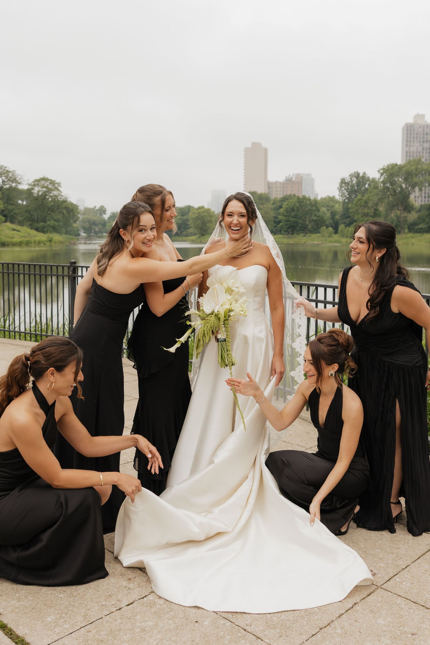 Bride in white gown laughs with five bridesmaids in black dresses near a lake with city skyline backdrop.