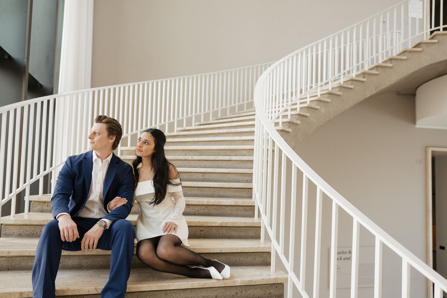 Couple sits together on elegant white staircase in formal attire for romantic engagement photos.