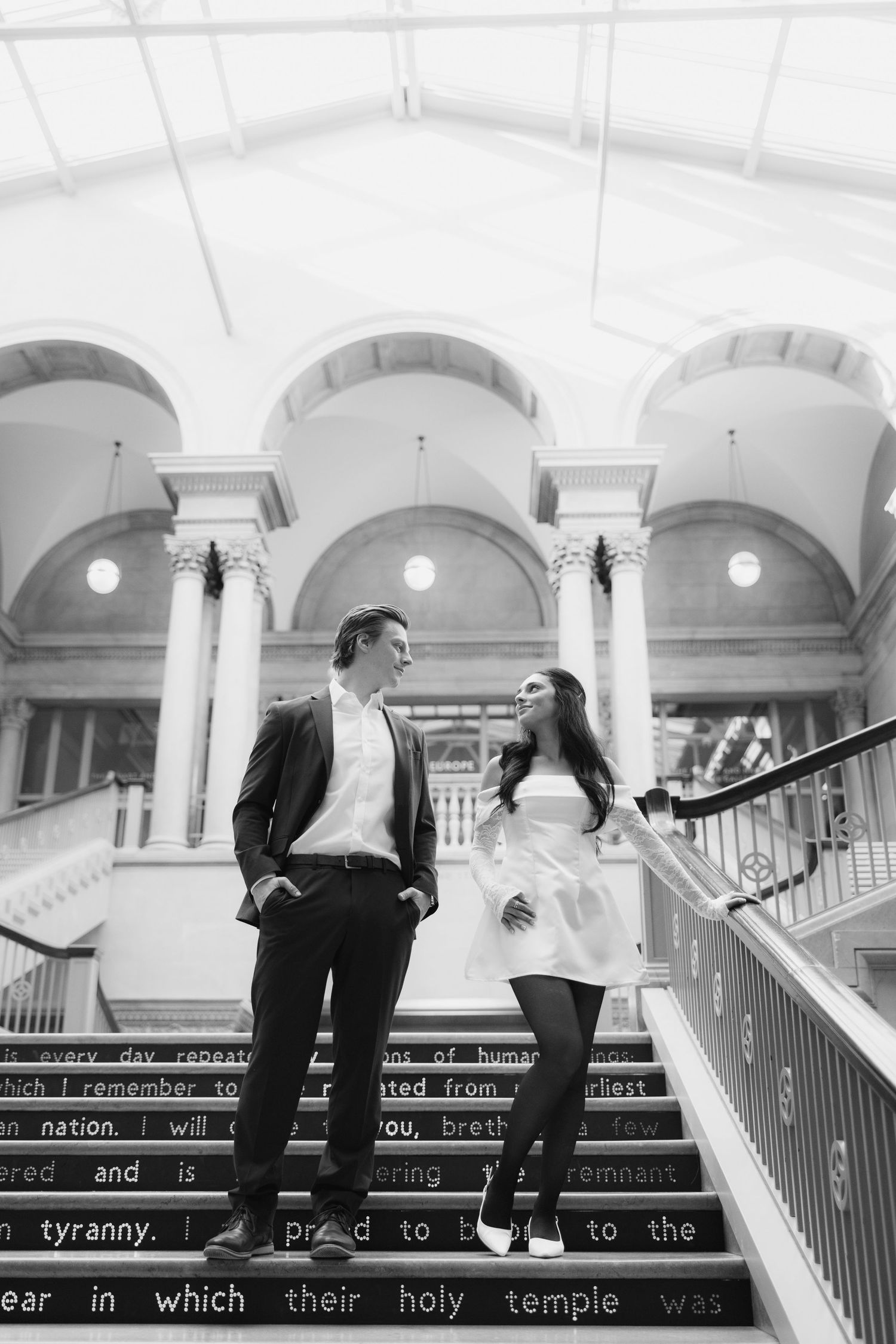 A couple stands on ornate marble steps with classical arched architecture in the background.