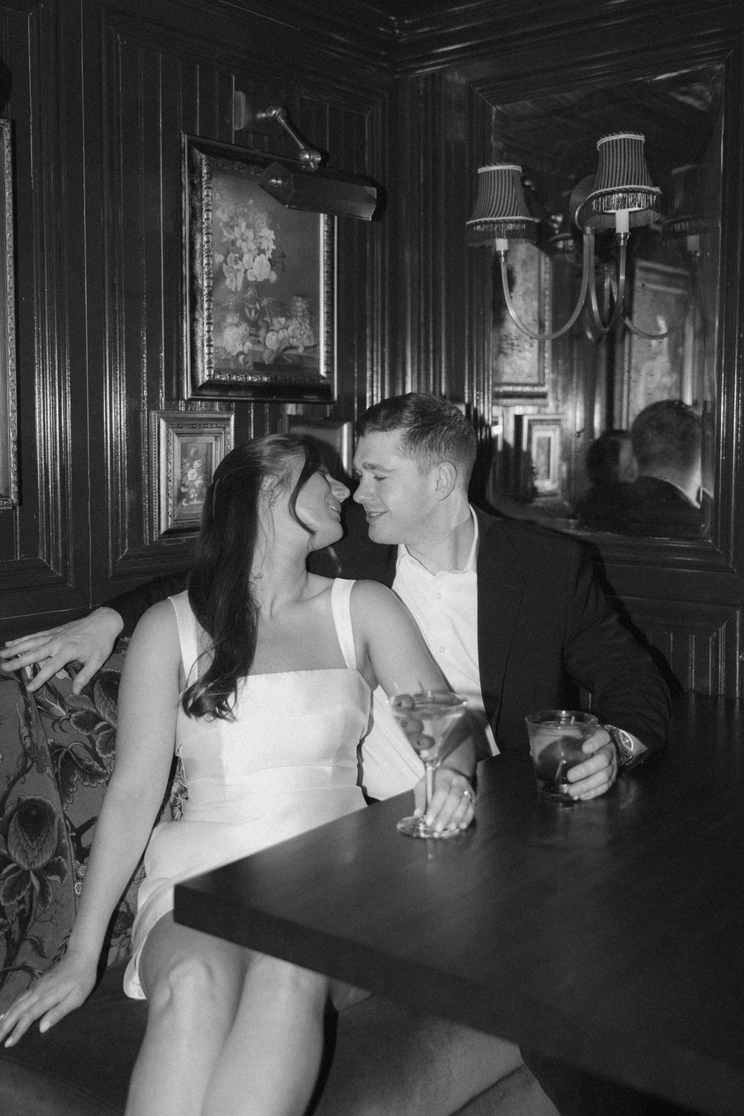 A couple in evening wear shares drinks and conversation in a dimly lit vintage bar setting.