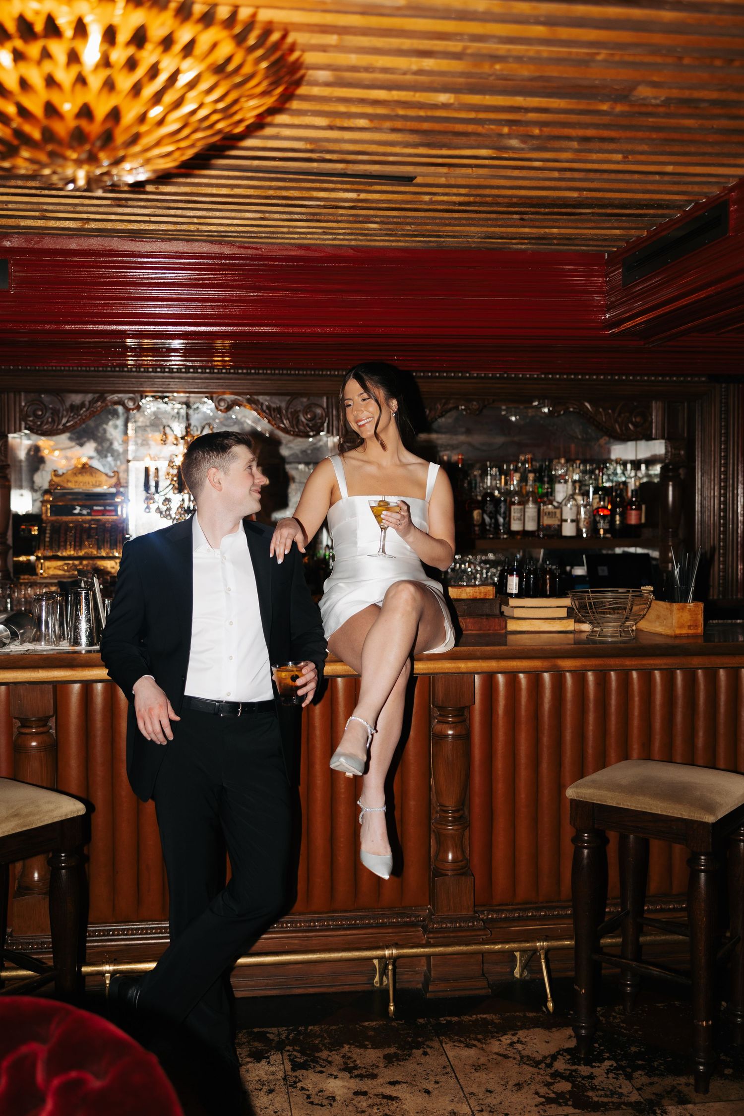 A couple in formal attire pose at an elegant vintage bar with wood paneling and decorative lighting.