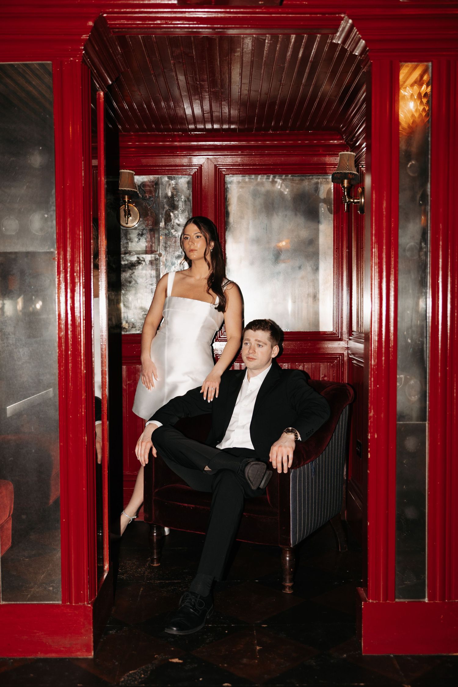 A couple poses together in an elegant red-framed booth at a dimly lit venue, wearing formal black and white attire.