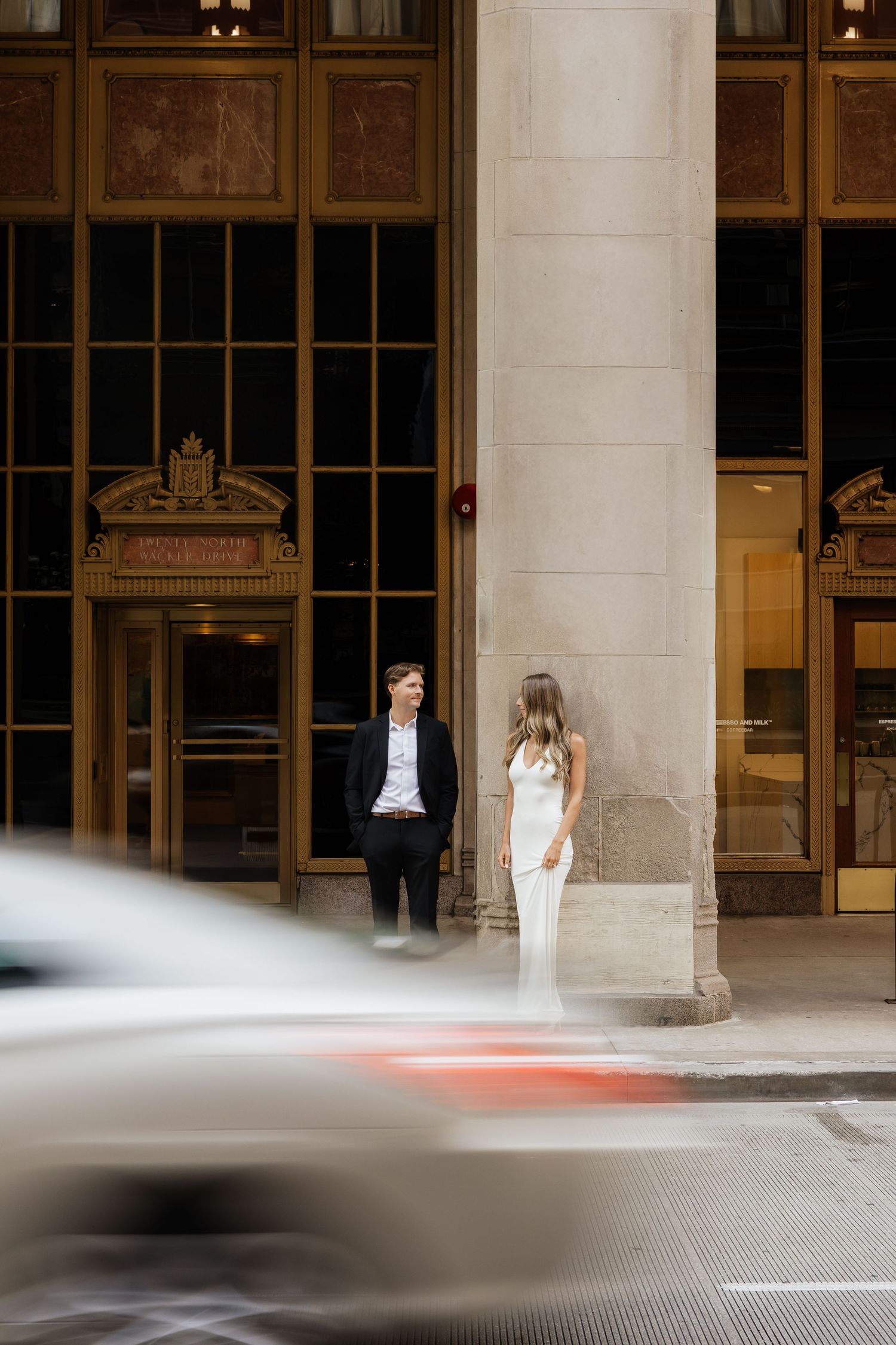 Couple in elegant attire standing outside a grand building entrance with a blurred car passing in the foreground.