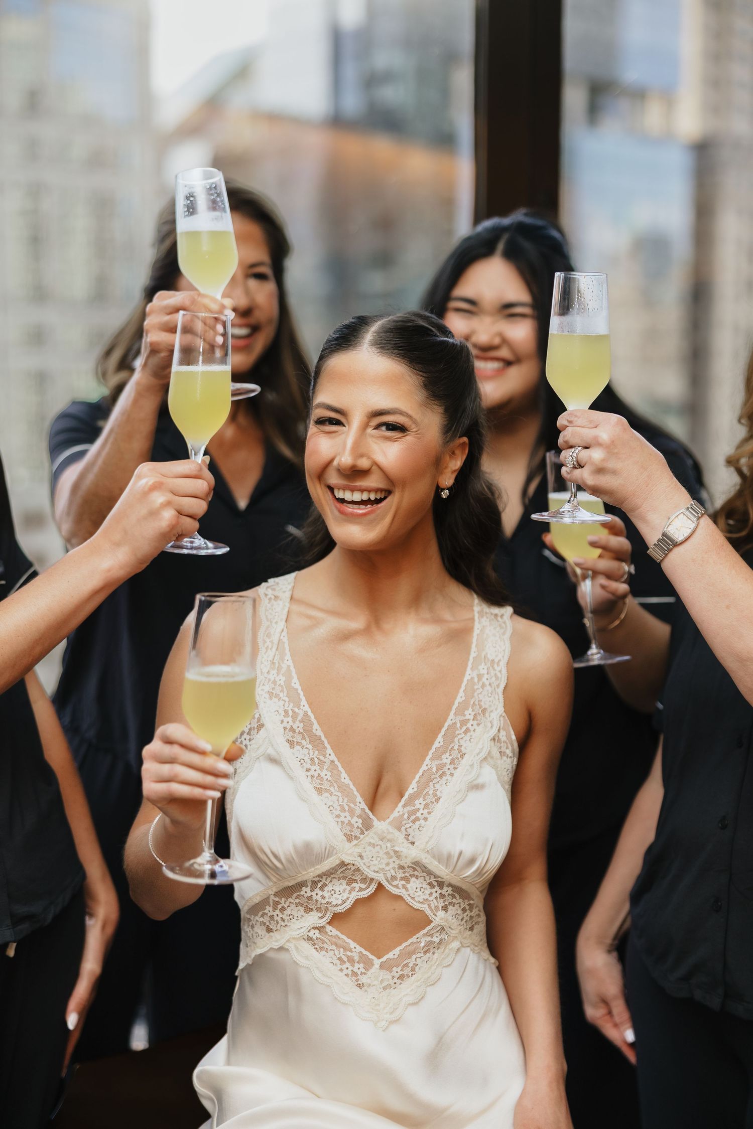 Bride in white lace gown toasts champagne with bridesmaids in black at a wedding celebration.