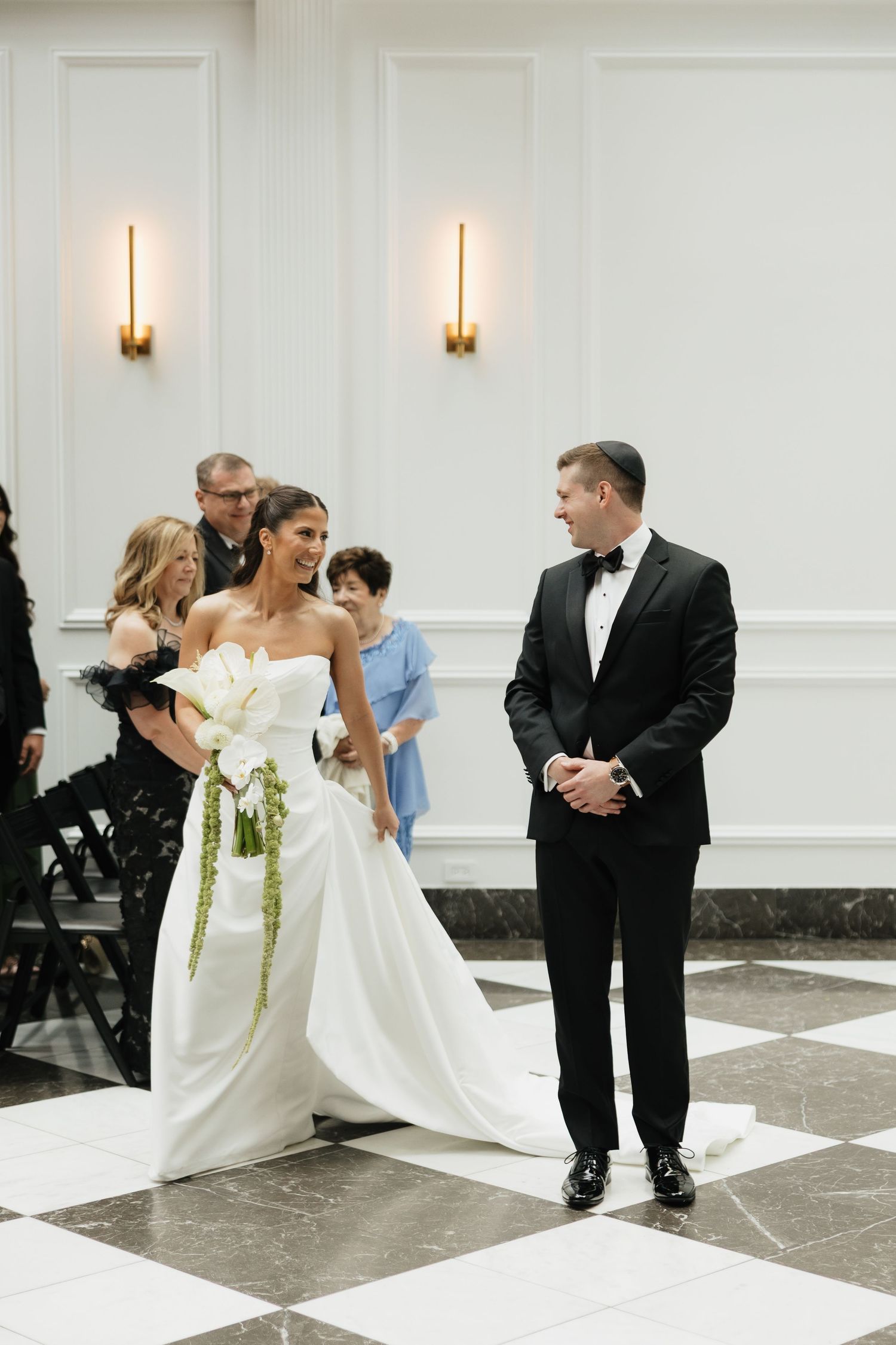 Radiant bride in strapless ball gown with floral bouquet walks toward smiling groom during indoor wedding ceremony.