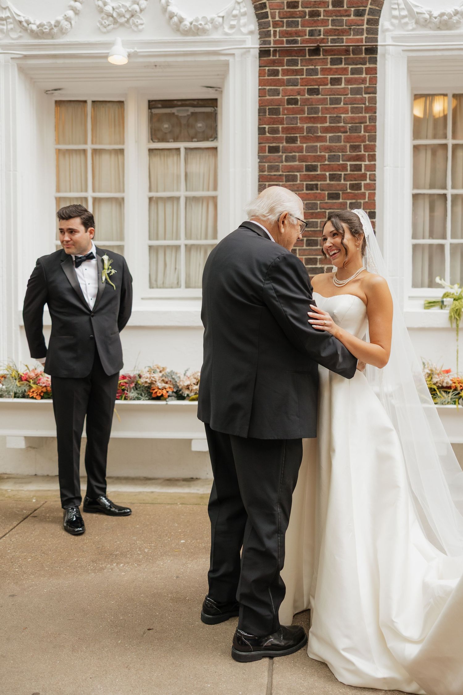 Bride in white gown smiling with older man while groom in tuxedo watches outside brick building.