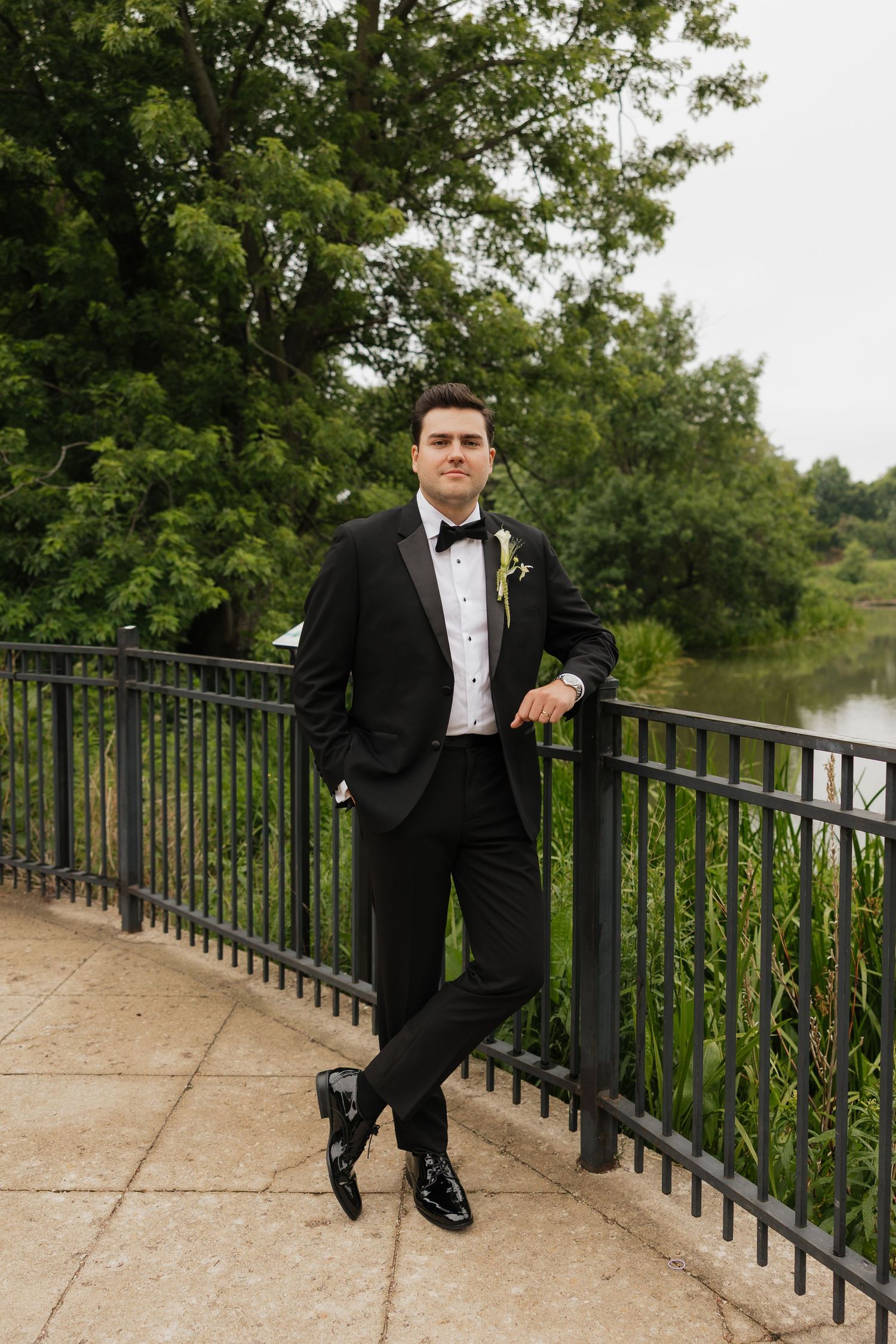 A groom in a black tuxedo with bow tie and boutonniere poses by iron railing near a serene pond.