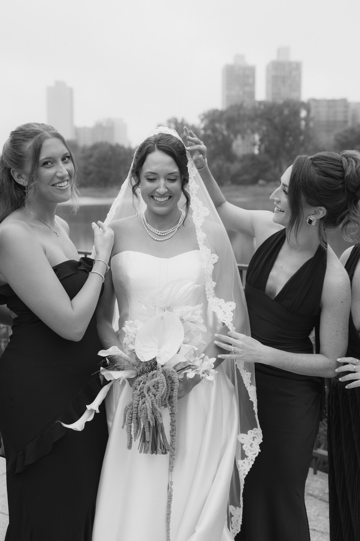 Black and white photo of a bride in a white gown and veil laughing with two bridesmaids in dark dresses outdoors.